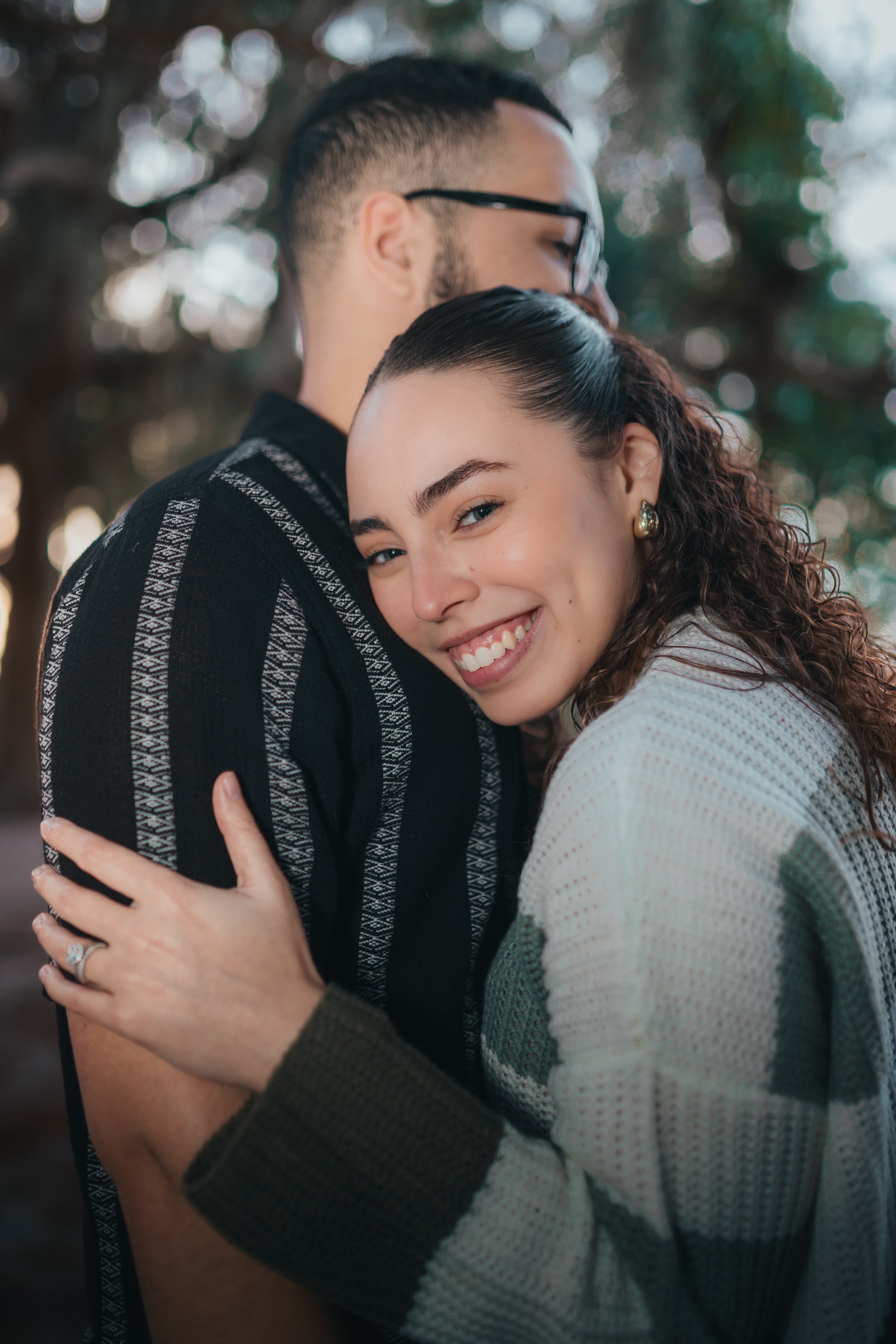 A smiling woman with curly hair hugging a man with glasses, outdoors with trees in the background.