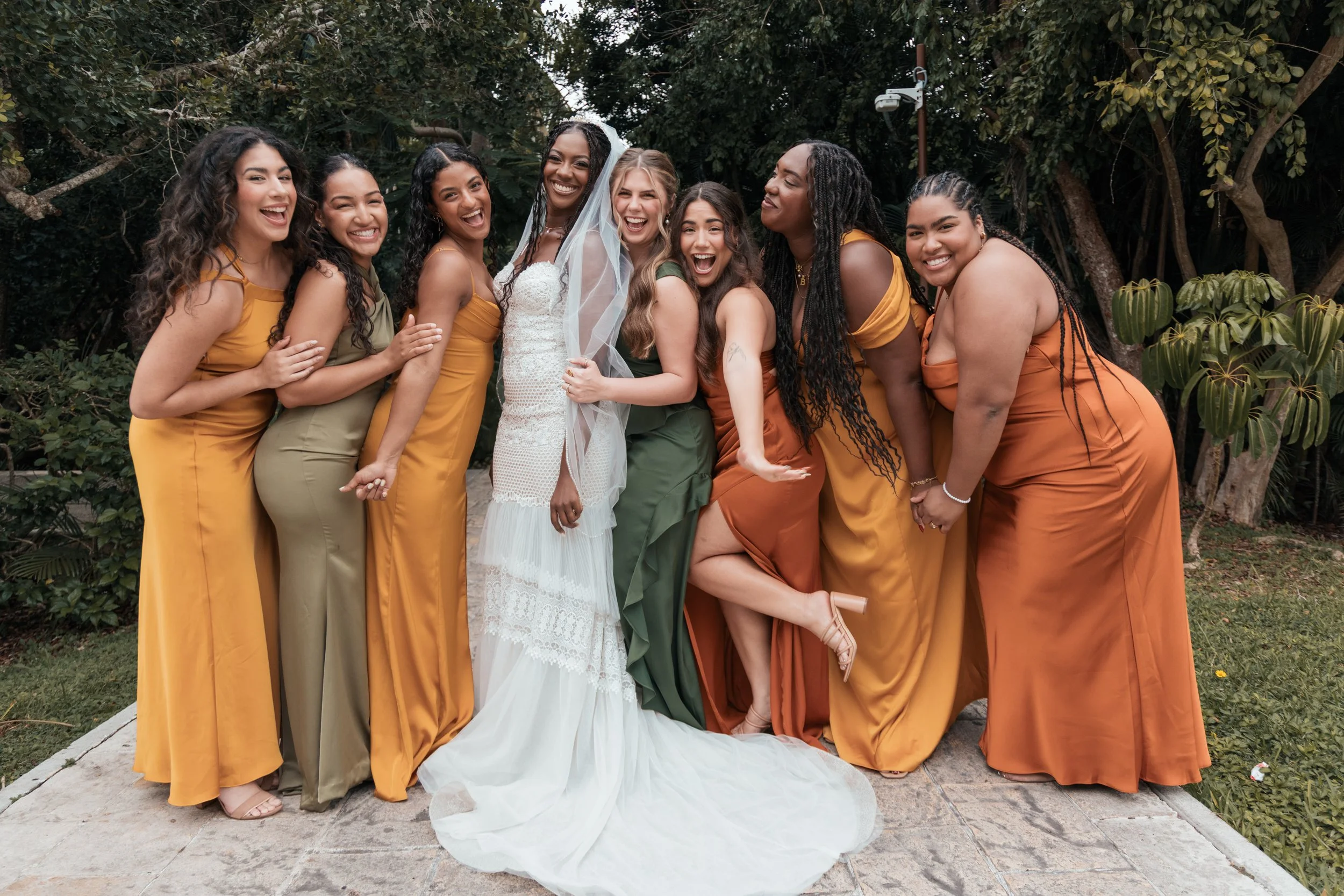 A group of nine women dressed in colorful gowns standing together outdoors, smiling and celebrating a wedding. The bride is in a white dress, wearing a veil, standing in the center. The others are in shades of yellow and green, embracing or posing pl