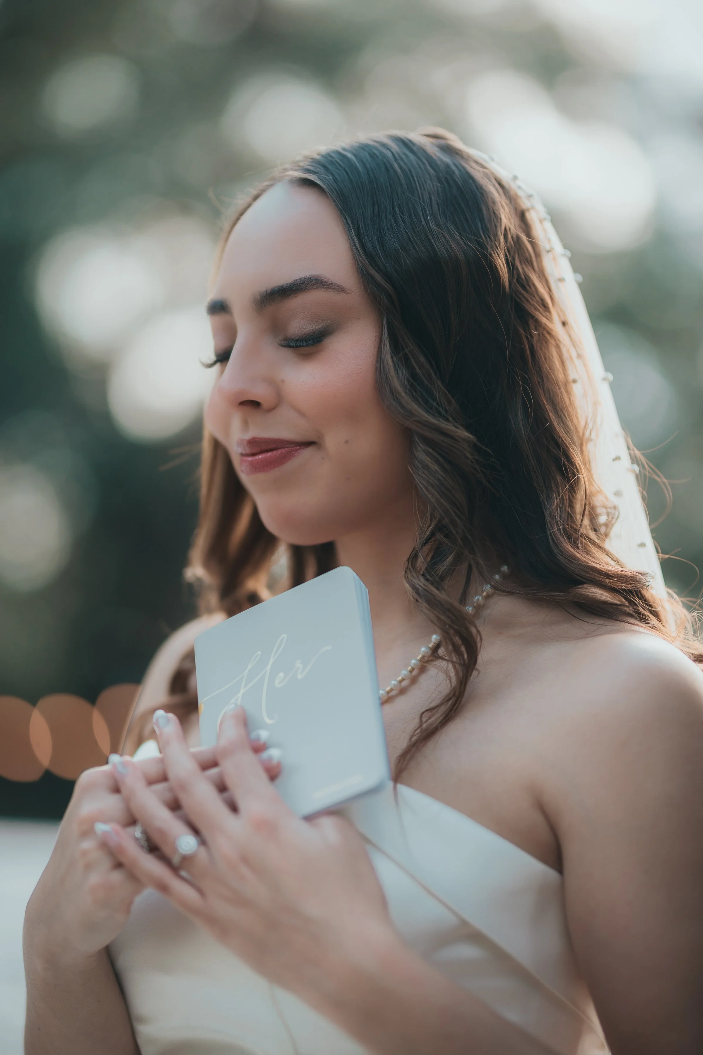 A bride with closed eyes, holding a small book titled "Her" close to her chest, outdoors with blurred lights and greenery in background.