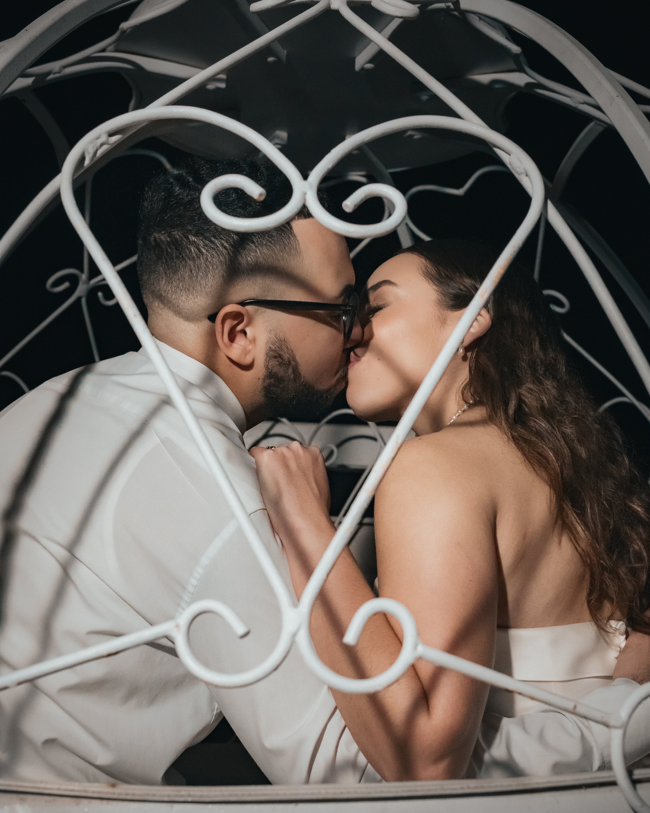 A couple sharing a kiss inside a decorative white metal gazebo with intricate scrollwork.
