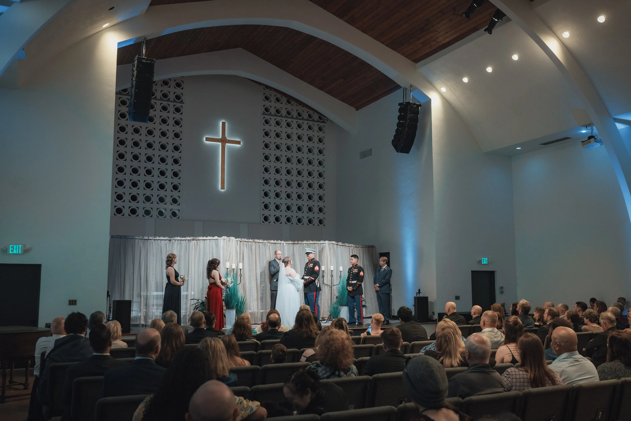 A wedding ceremony taking place in a church with a large illuminated cross on the wall, a bride and groom exchanging vows, with bridesmaids and groomsmen standing nearby, and guests seated facing the altar.