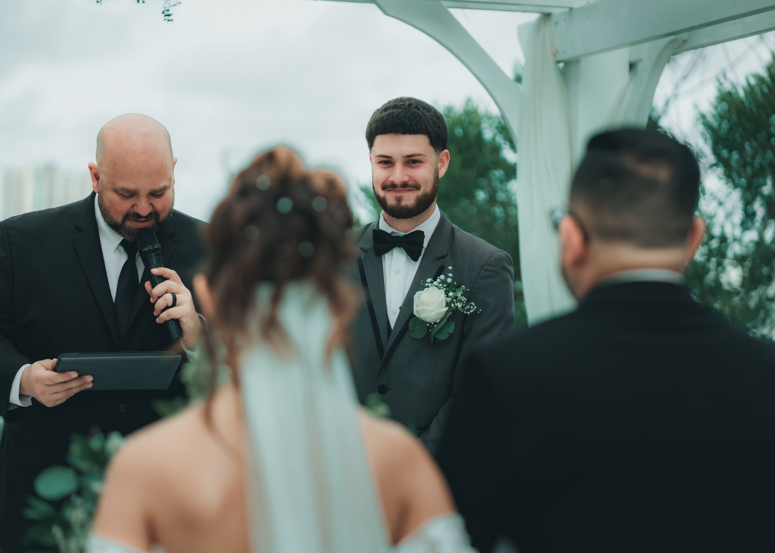 A groom in a tuxedo and bow tie smiling during a wedding ceremony, standing under a white canopy, with an officiant holding a microphone and a tablet, and the bride and another man with their backs turned in the foreground.