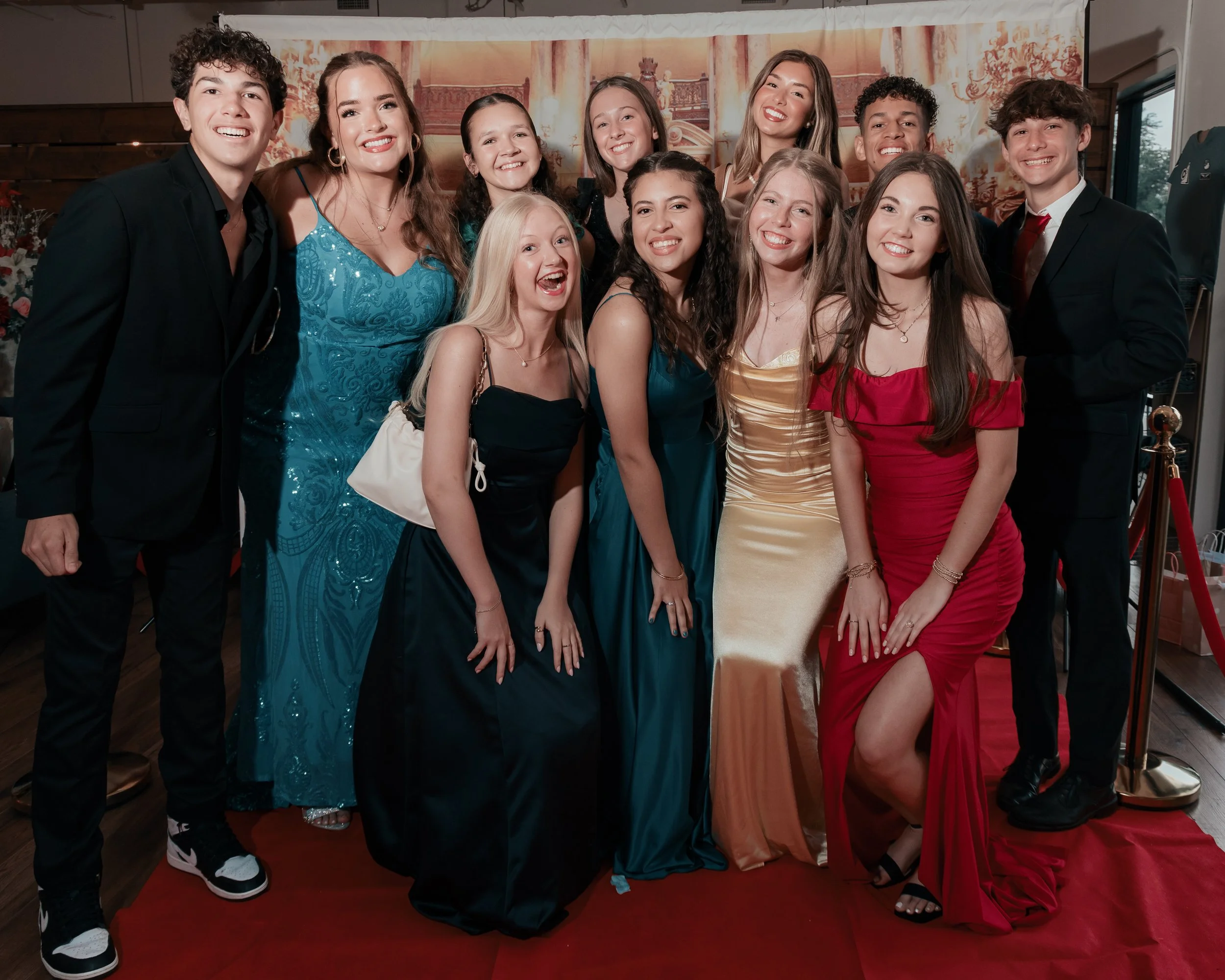 A group of fifteen young women and men dressed in formal attire, smiling and posing on a red carpet at an indoor event.