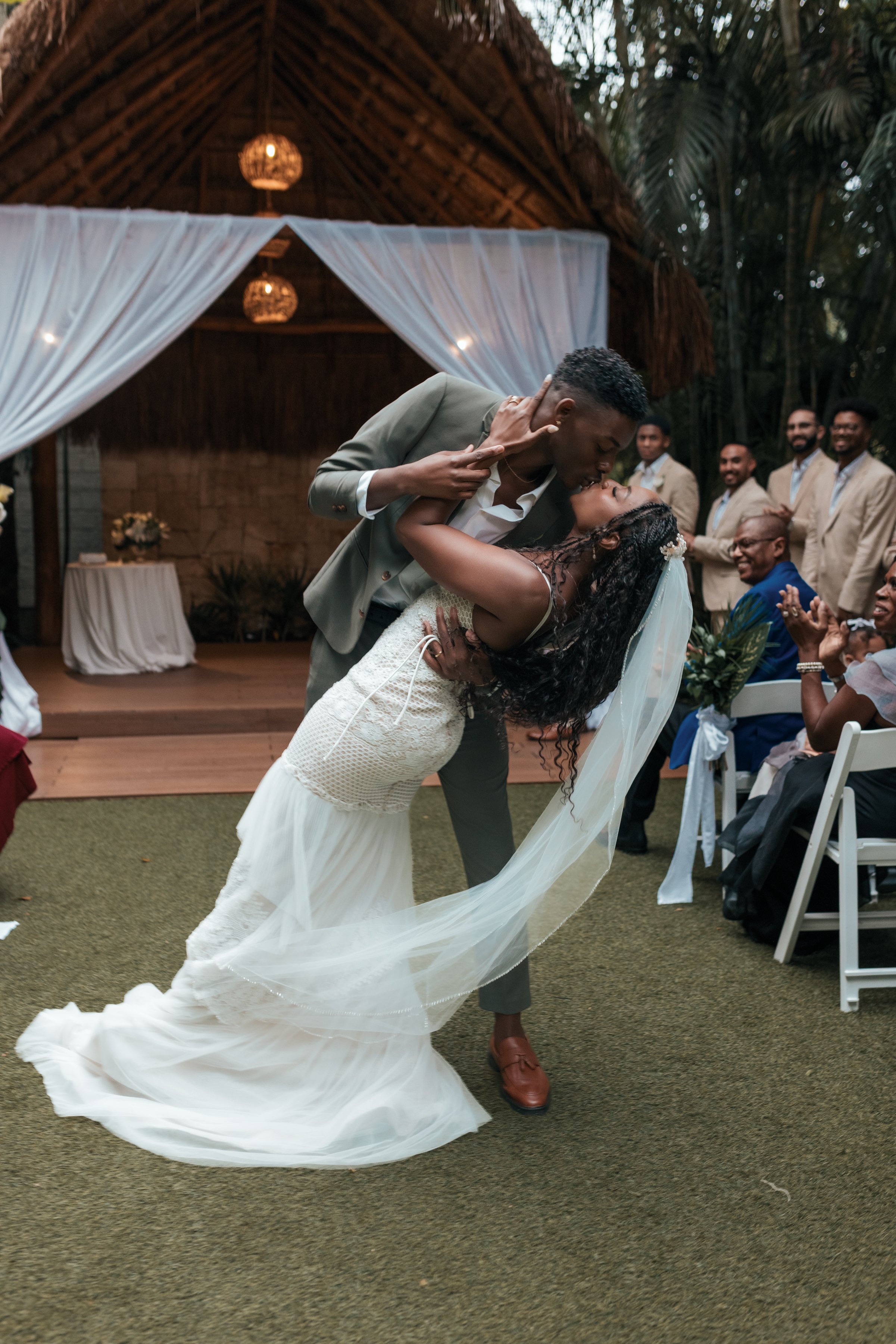 A bride and groom sharing a kiss during their wedding reception. The bride is wearing a white gown and a veil, and the groom is in a gray suit. Guests are seated and standing around, clapping and smiling.