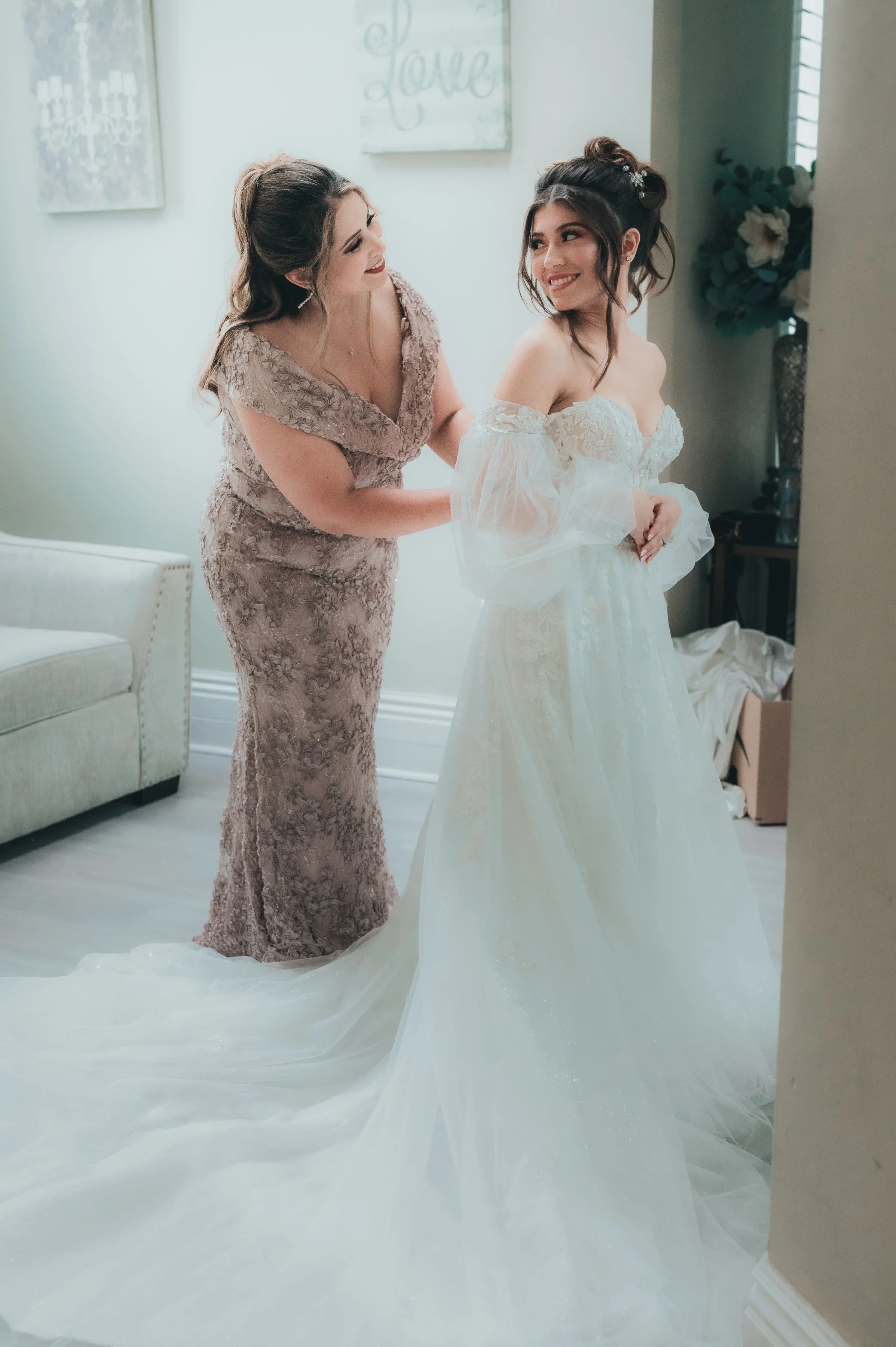 A bride in a white wedding gown smiling as an older woman helps her with her dress in a room with decorative wall art.