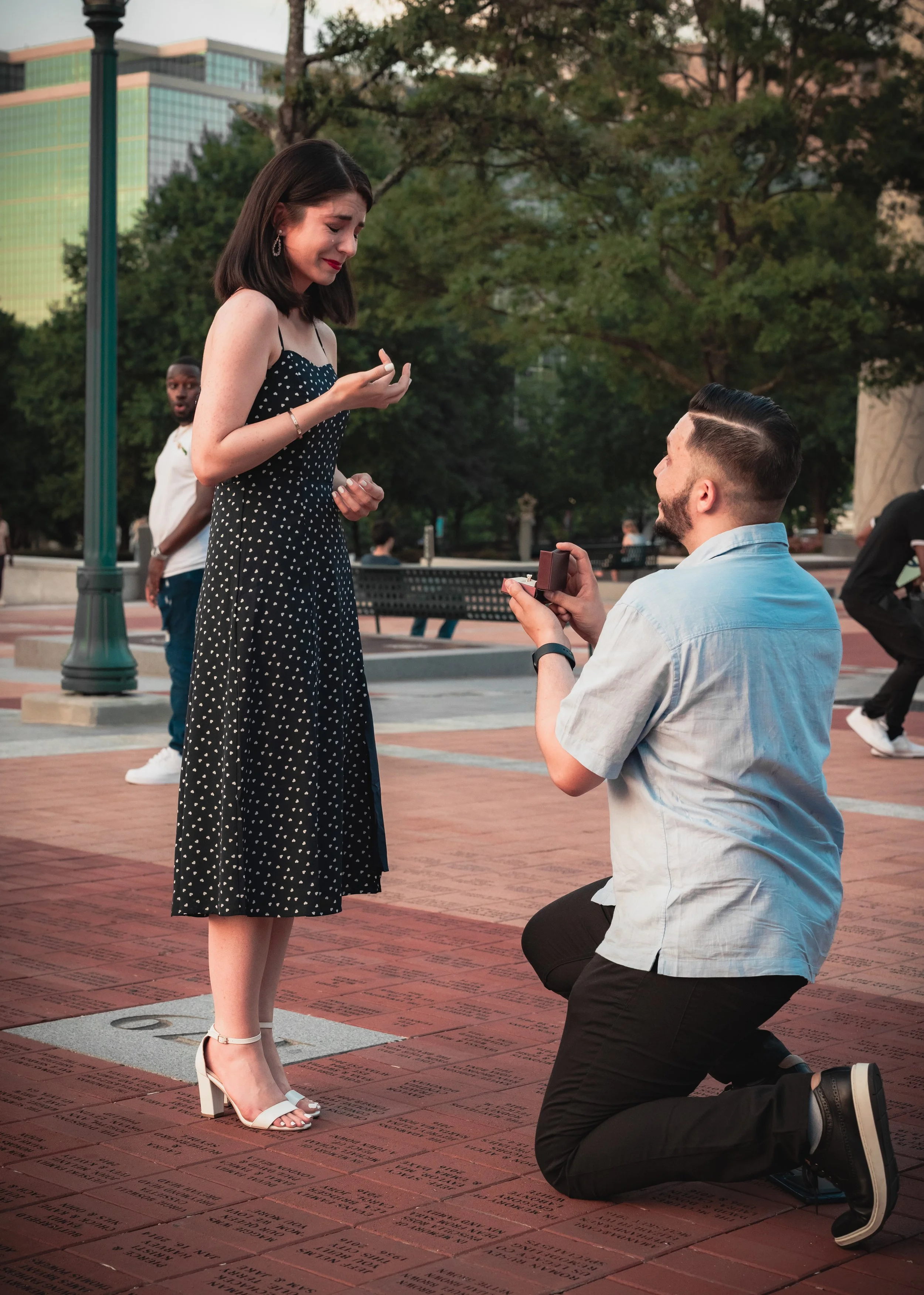 A man proposing marriage with a ring box to a woman in a black dress with white polka dots at an outdoor park during sunset as onlookers watch.
