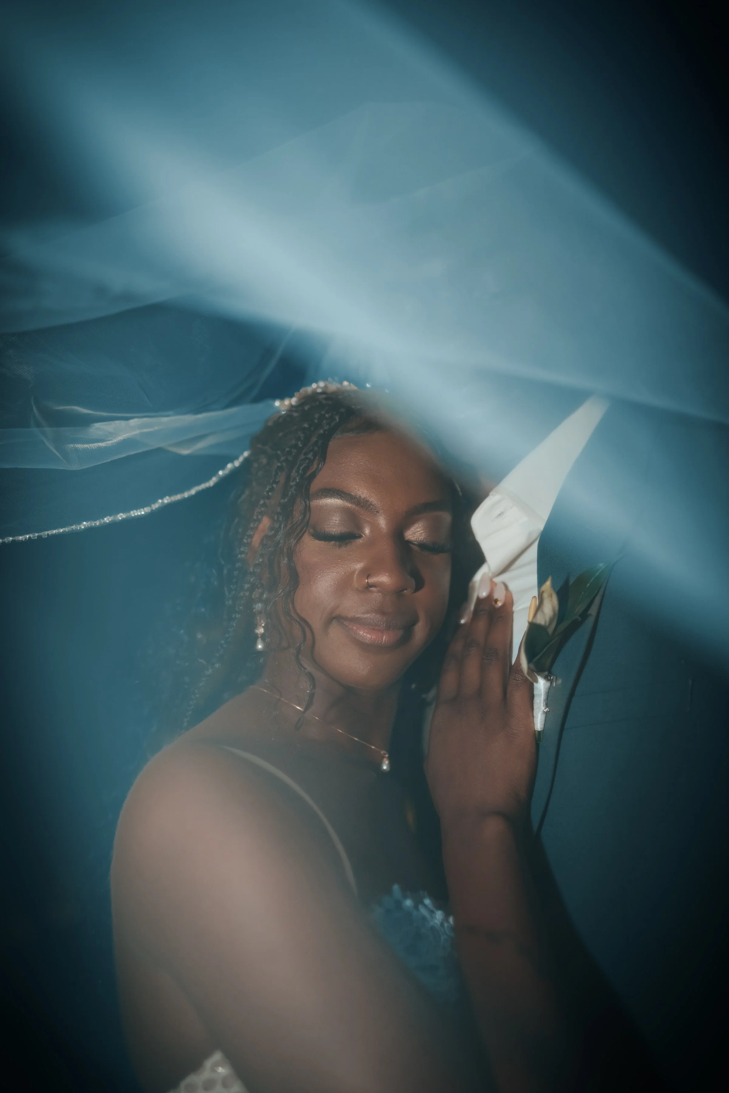A woman with dark skin and braided hair, dressed in a white wedding gown, peacefully resting her head and hand on a man's shoulder, who is wearing a dark suit. Soft blue lighting and sheer fabric create a dreamy atmosphere.