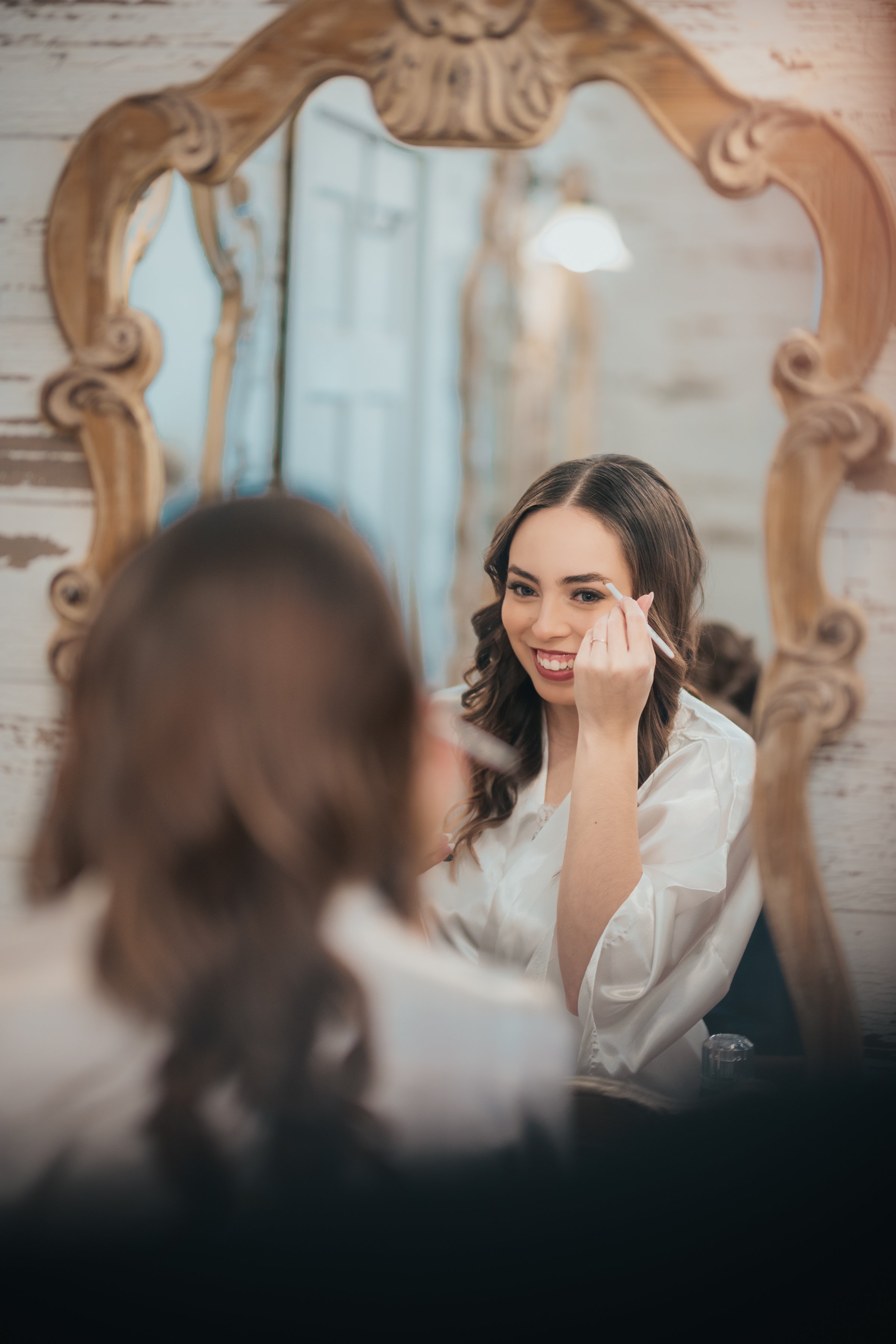 A woman with long, wavy brown hair applying makeup in front of a mirror with a decorative wooden frame.