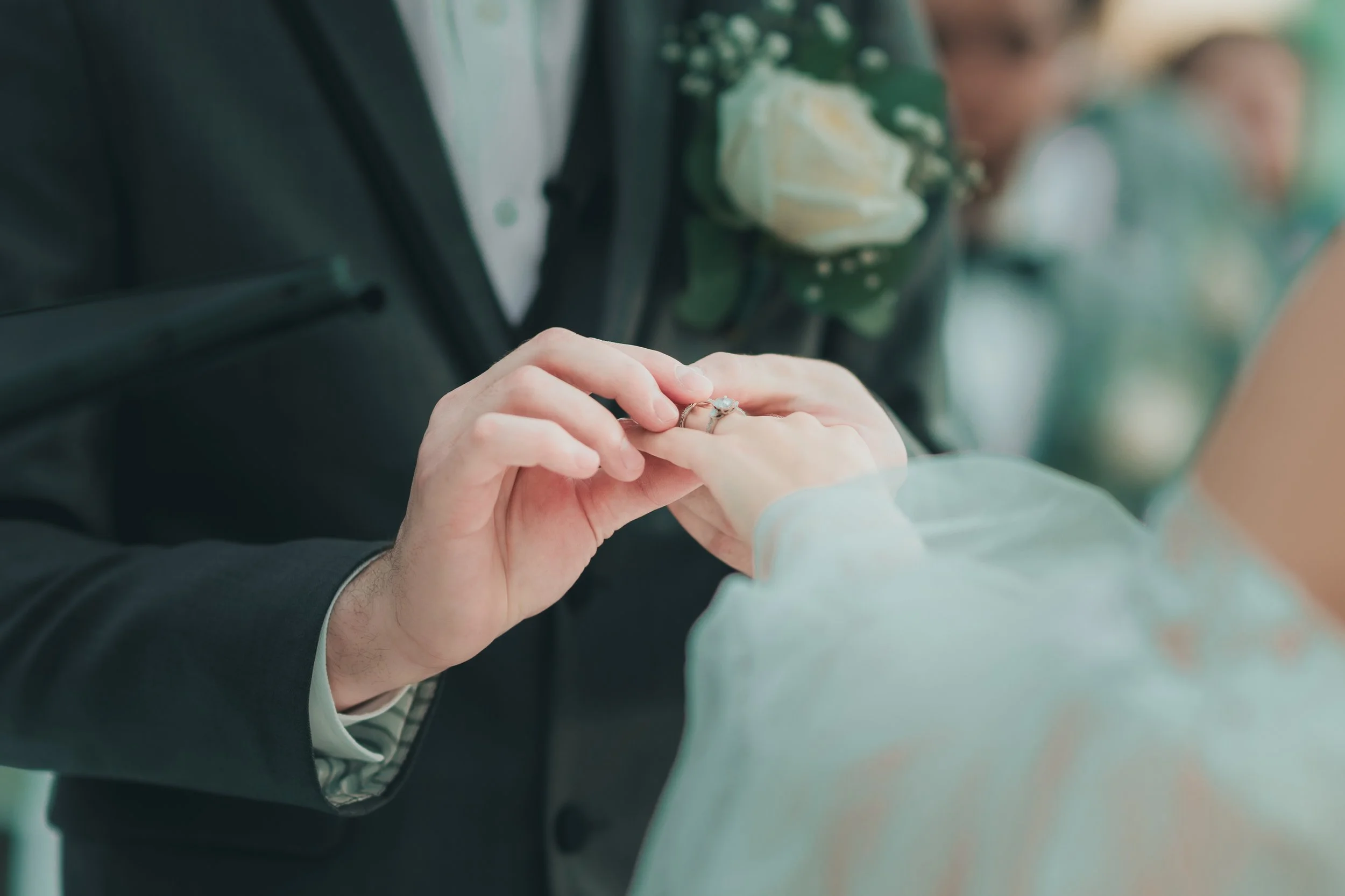 A person in a wedding dress placing a ring on a groom's finger, who is wearing a black suit with a boutonniere.
