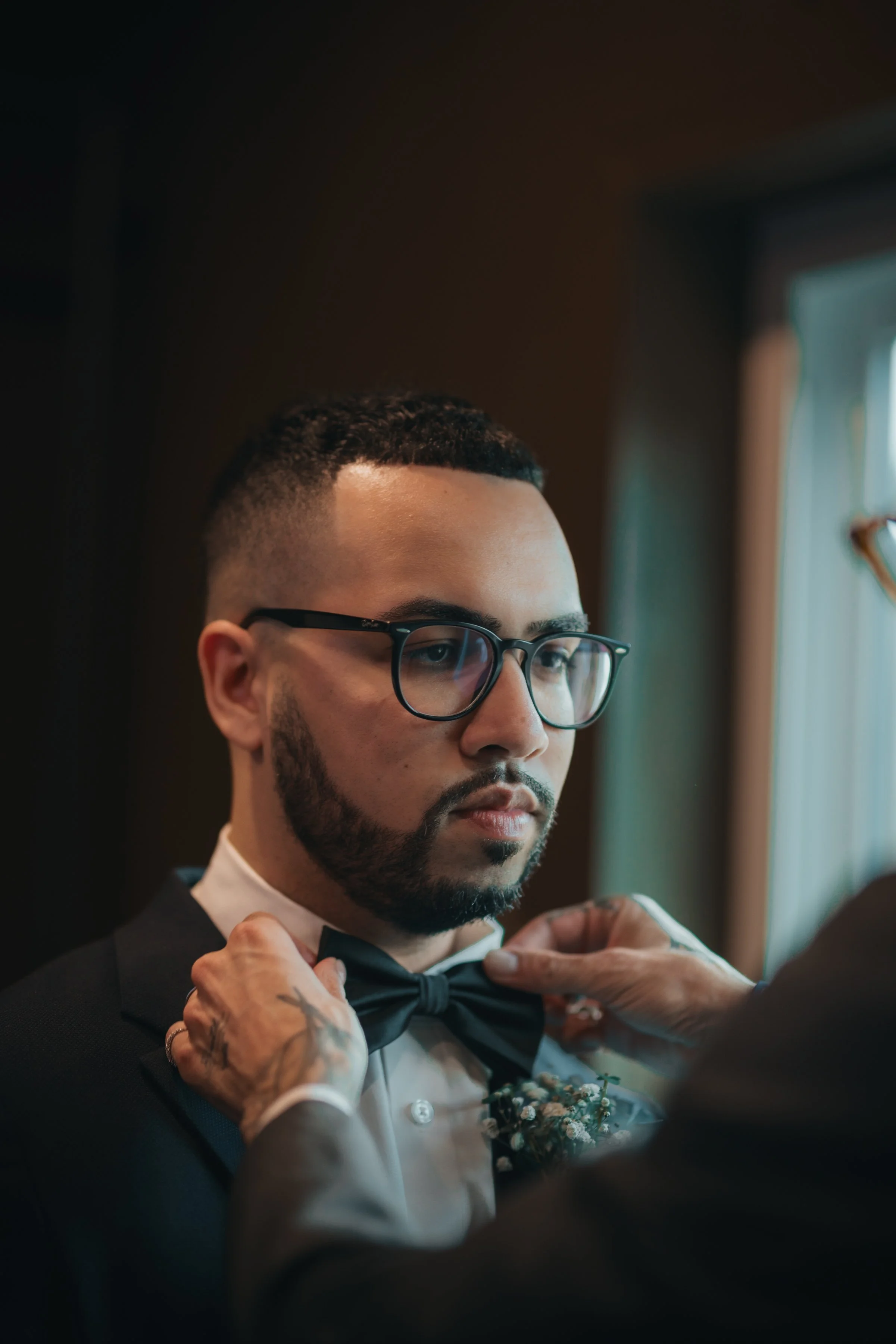 A man with glasses, a beard, and short hair is being helped with a bow tie, wearing a tuxedo with a boutonniere, by another person.