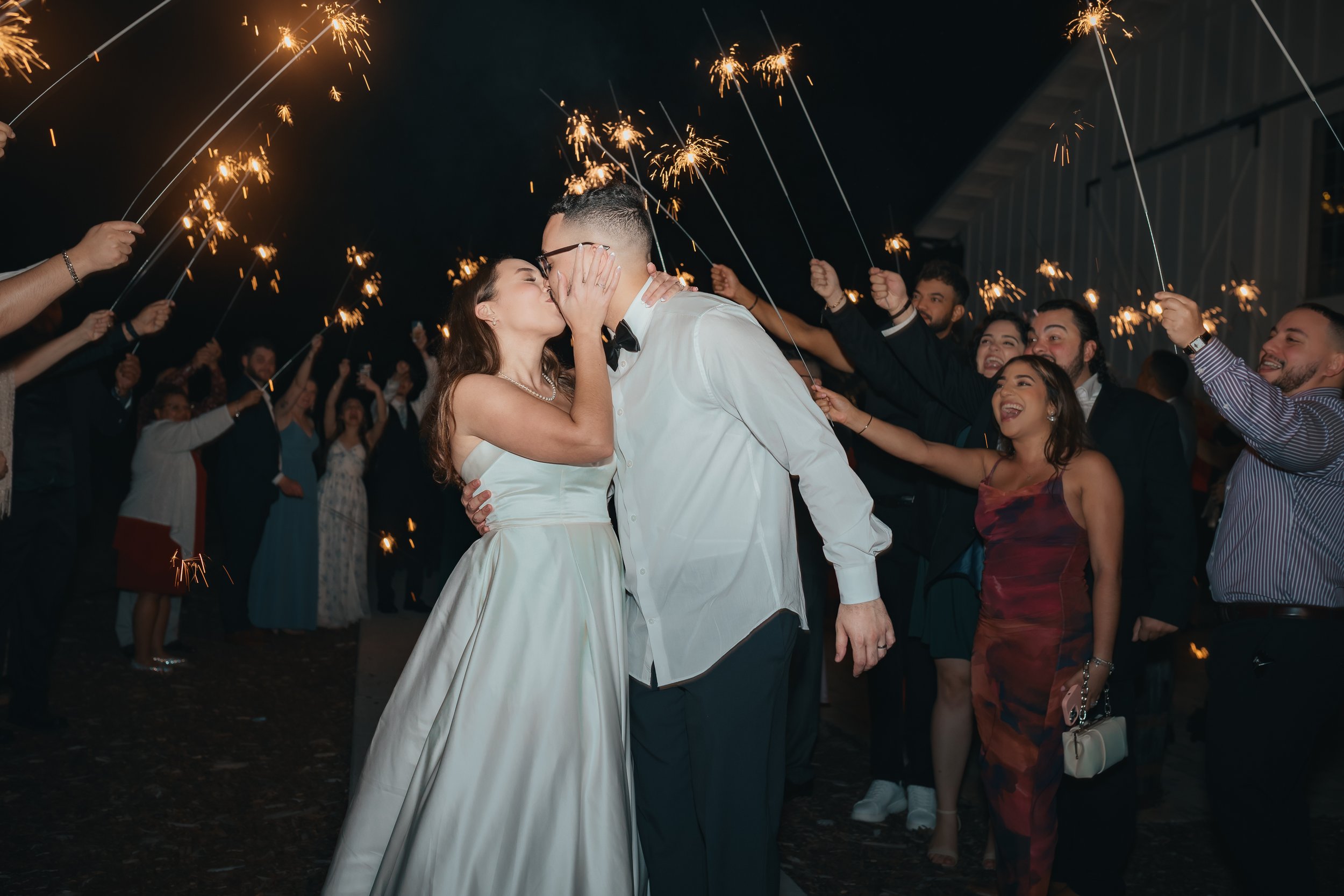 A newlywed couple shares a kiss at night during their wedding celebration surrounded by guests holding sparklers.