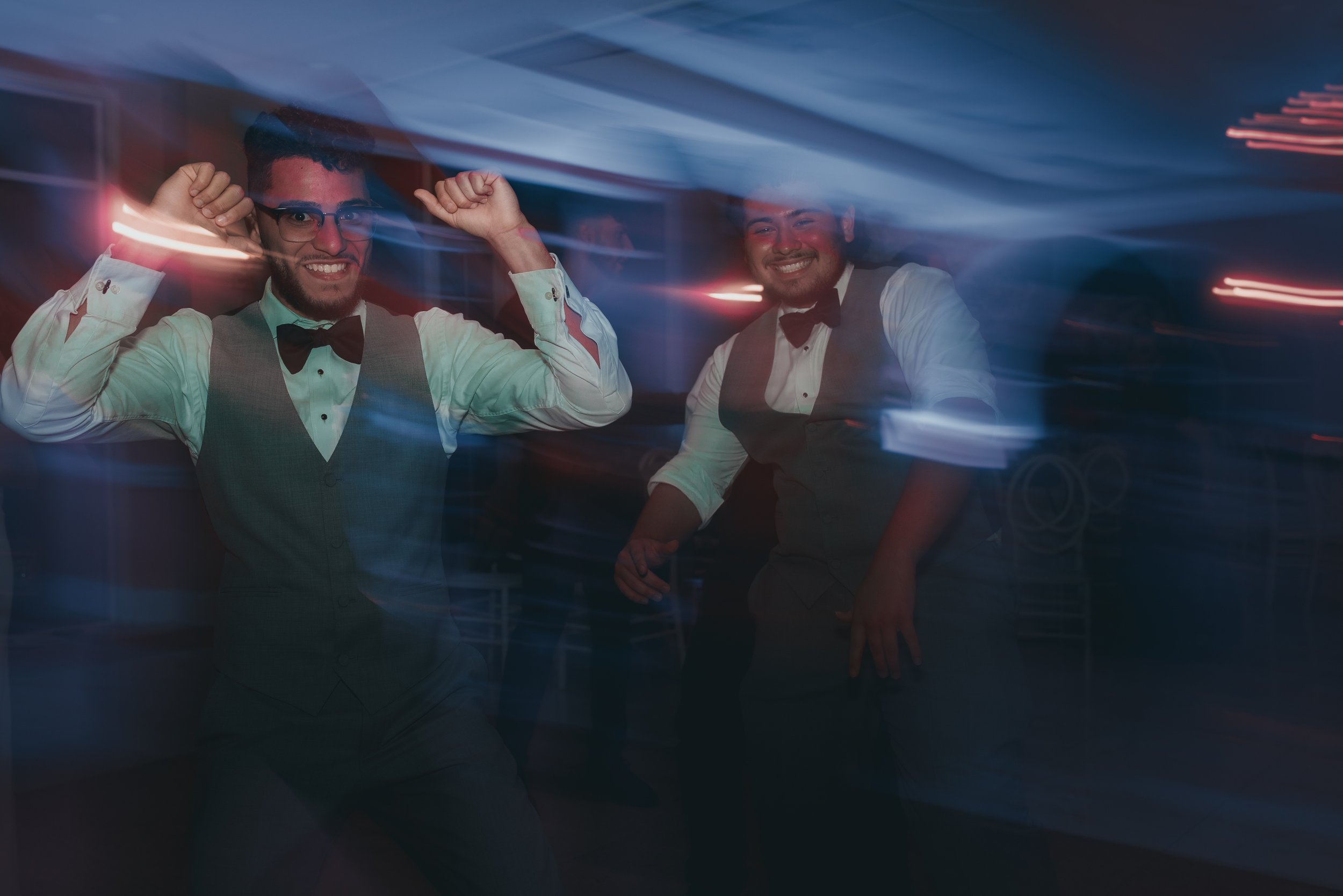 Two men in formal attire dancing at a party with colorful lights and motion blur effects.
