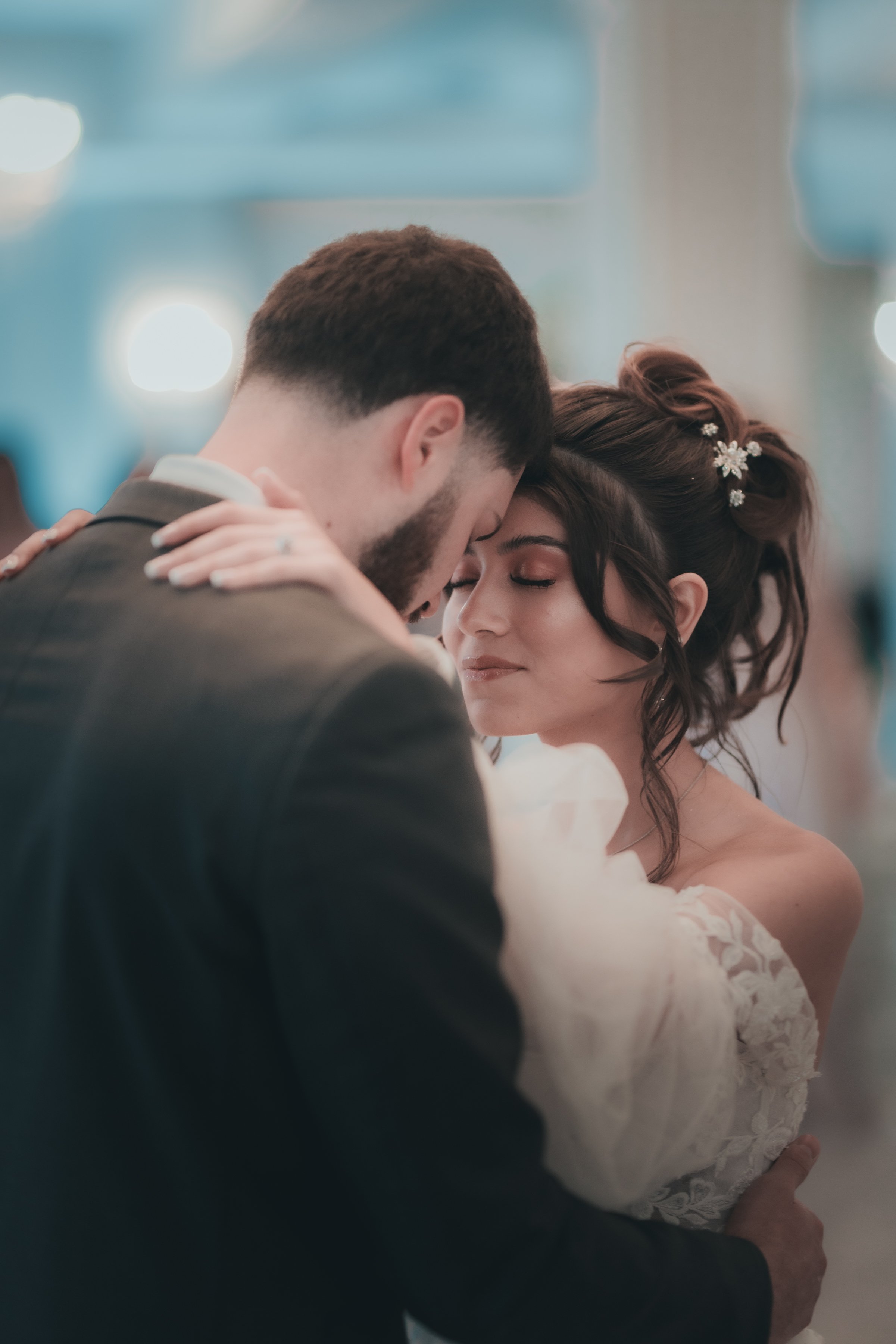 A bride and groom sharing a close, intimate dance at their wedding reception, eyes closed, with soft lighting and a blurred background.