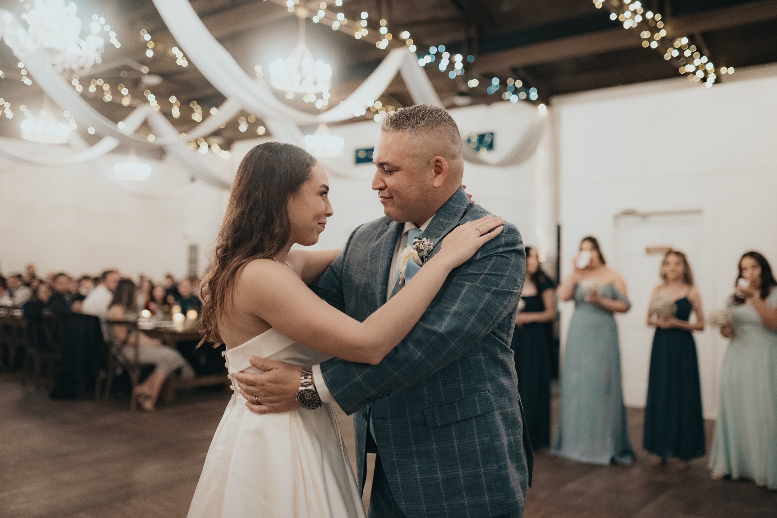 A bride and groom sharing their first dance at a wedding reception, with guests watching in the background and elegant drapery and string lights decorating the ceiling.
