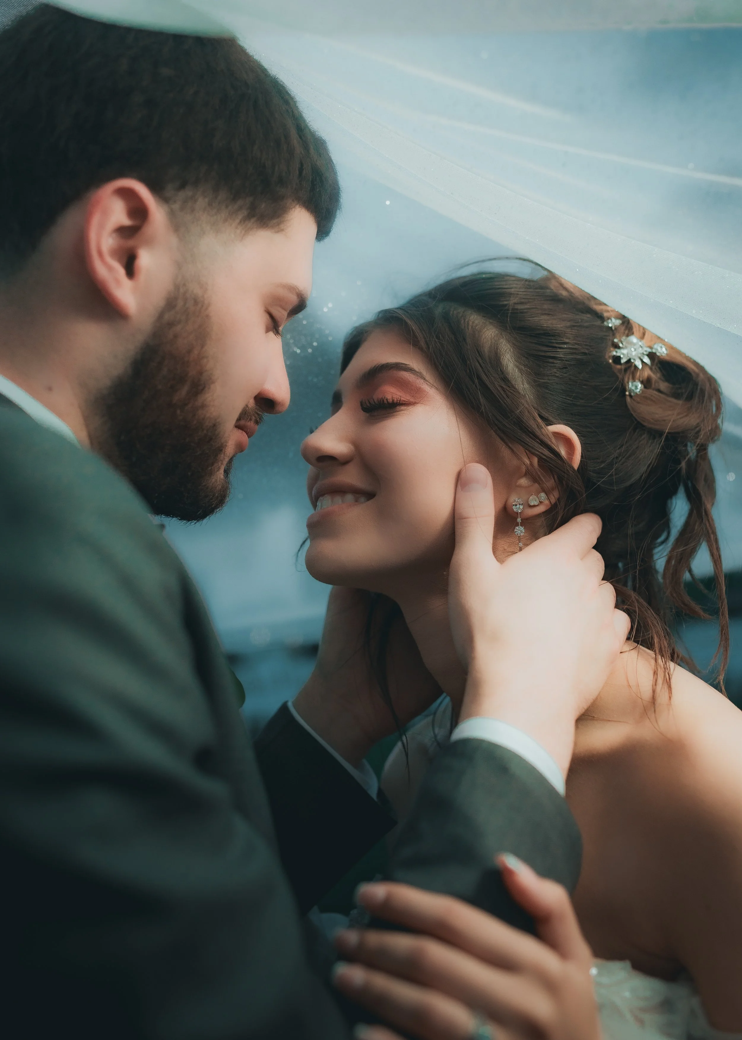A couple in wedding attire embraces closely inside a car, with the man gently holding the woman's face and the woman smiling with her eyes closed, showing affection and happiness.