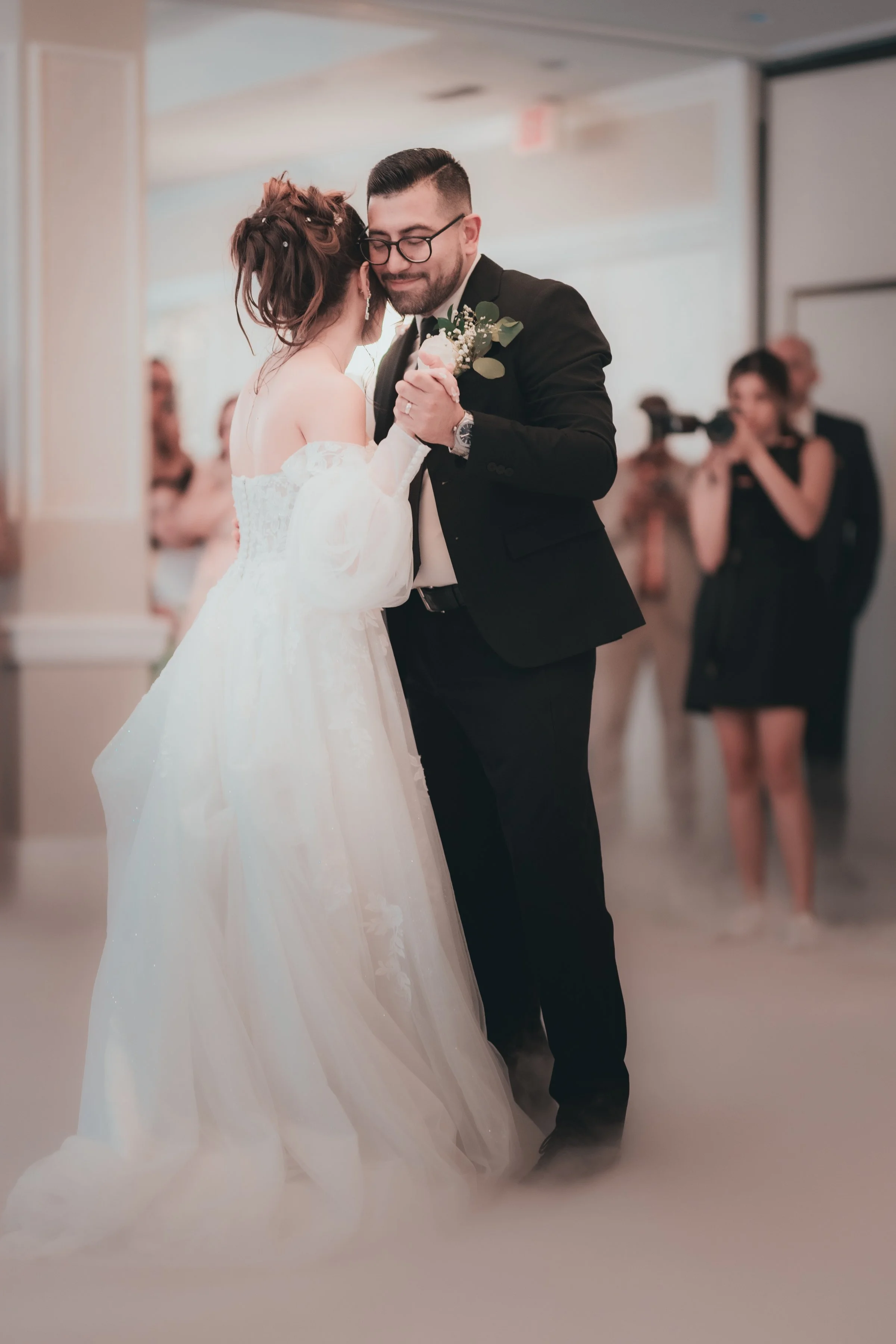 A bride and groom sharing a dance at their wedding reception, with the bride in a white wedding gown and the groom in a black suit, with a woman with a camera in the background.