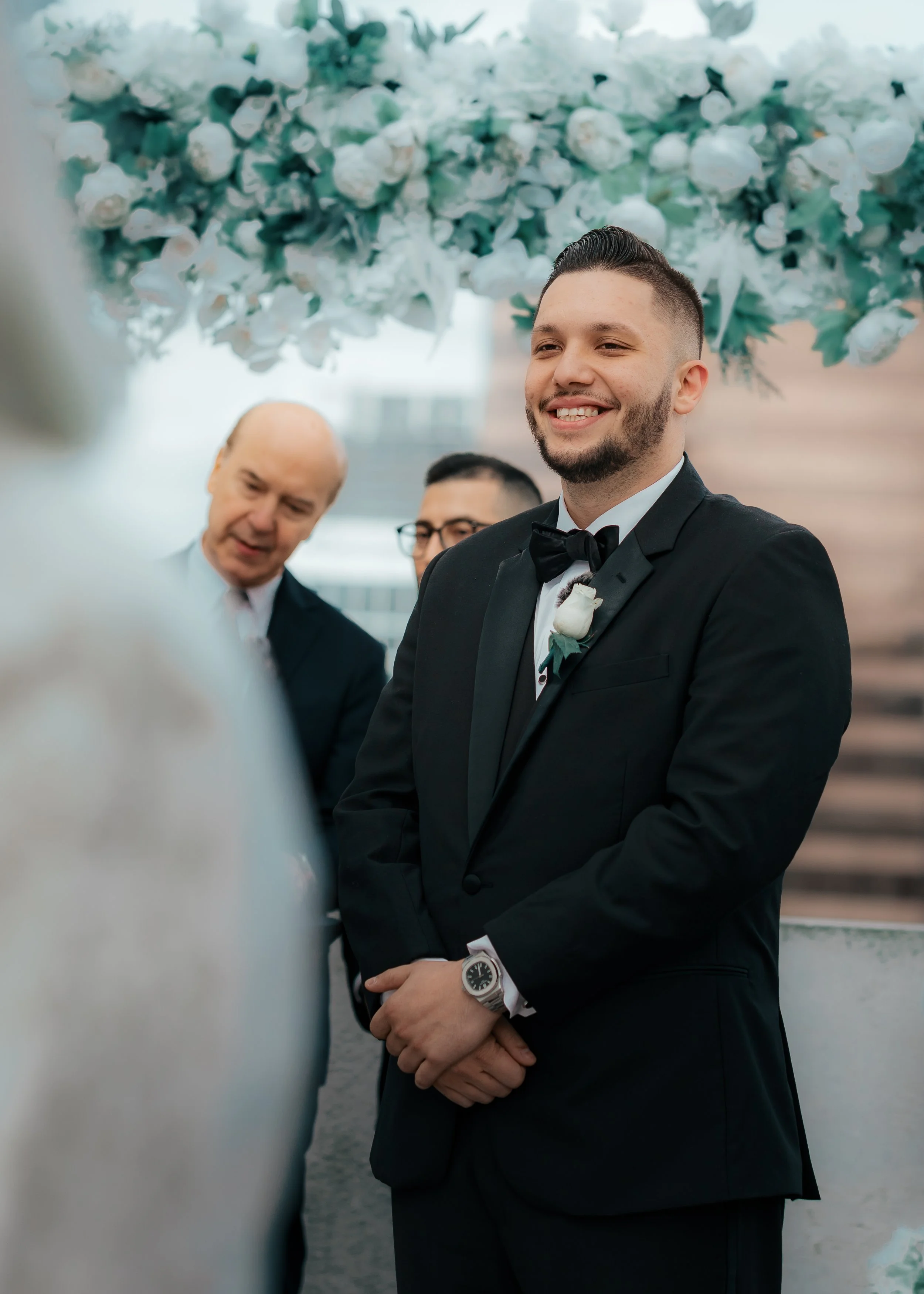 A groom standing in a black tuxedo with a bow tie, smiling during his wedding ceremony, with an officiant and another man in the background, and a floral arrangement overhead.