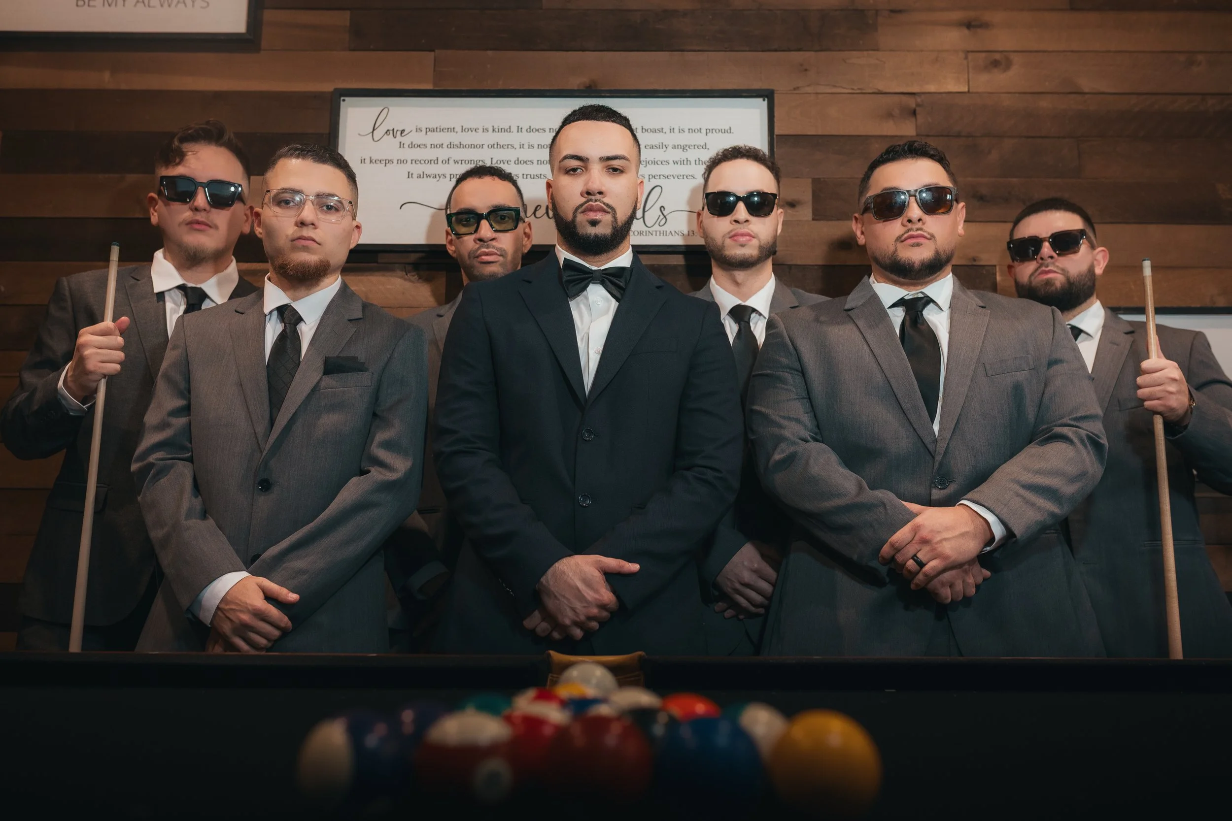 A group of seven men dressed in formal suits and sunglasses pose in front of a wood-paneled wall, with two holding pool cues, behind a pool table with billiard balls.