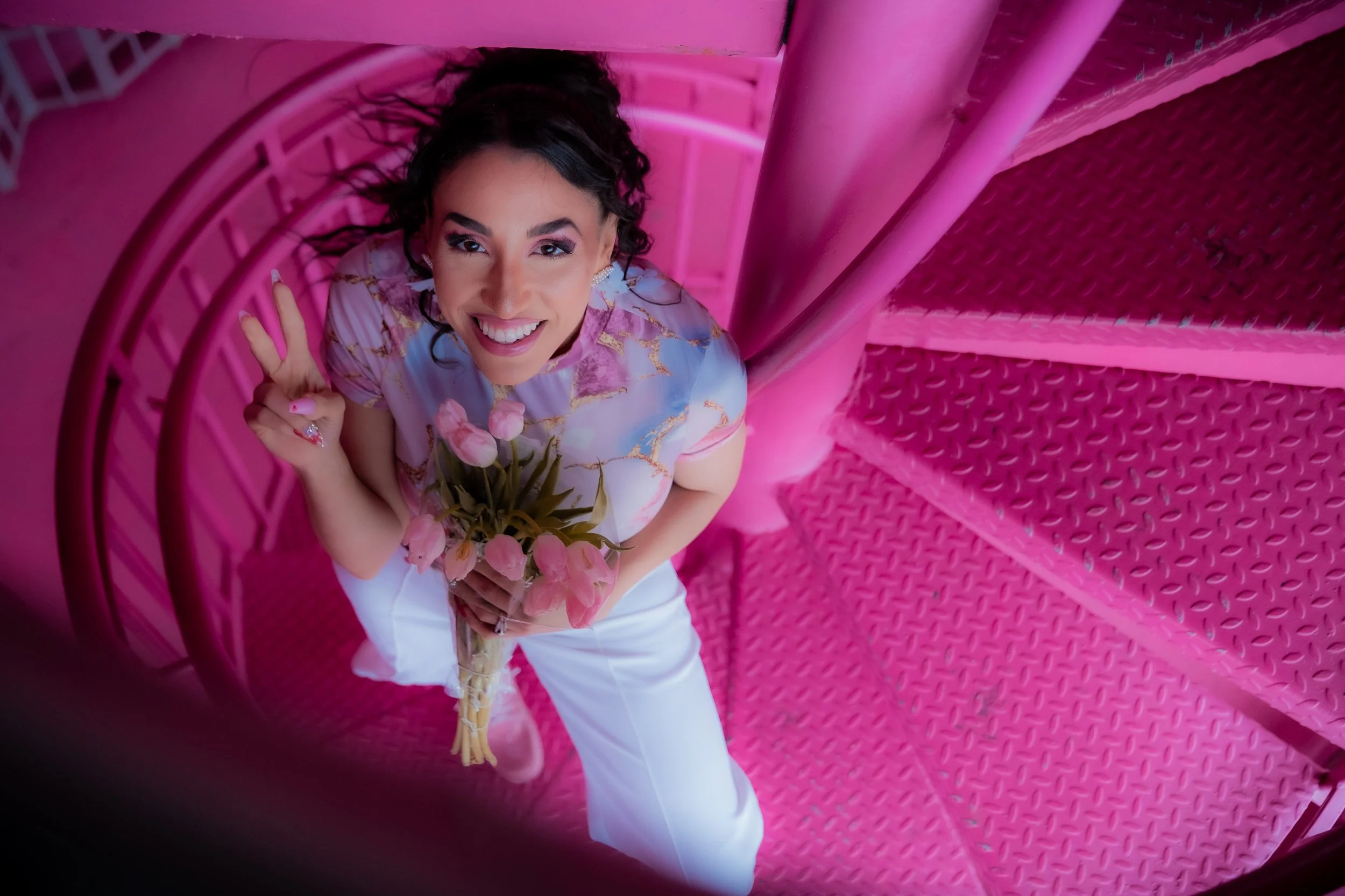 A smiling woman with dark curly hair, holding a bouquet of pink flowers, standing on pink metal spiral staircase and flashing a peace sign.