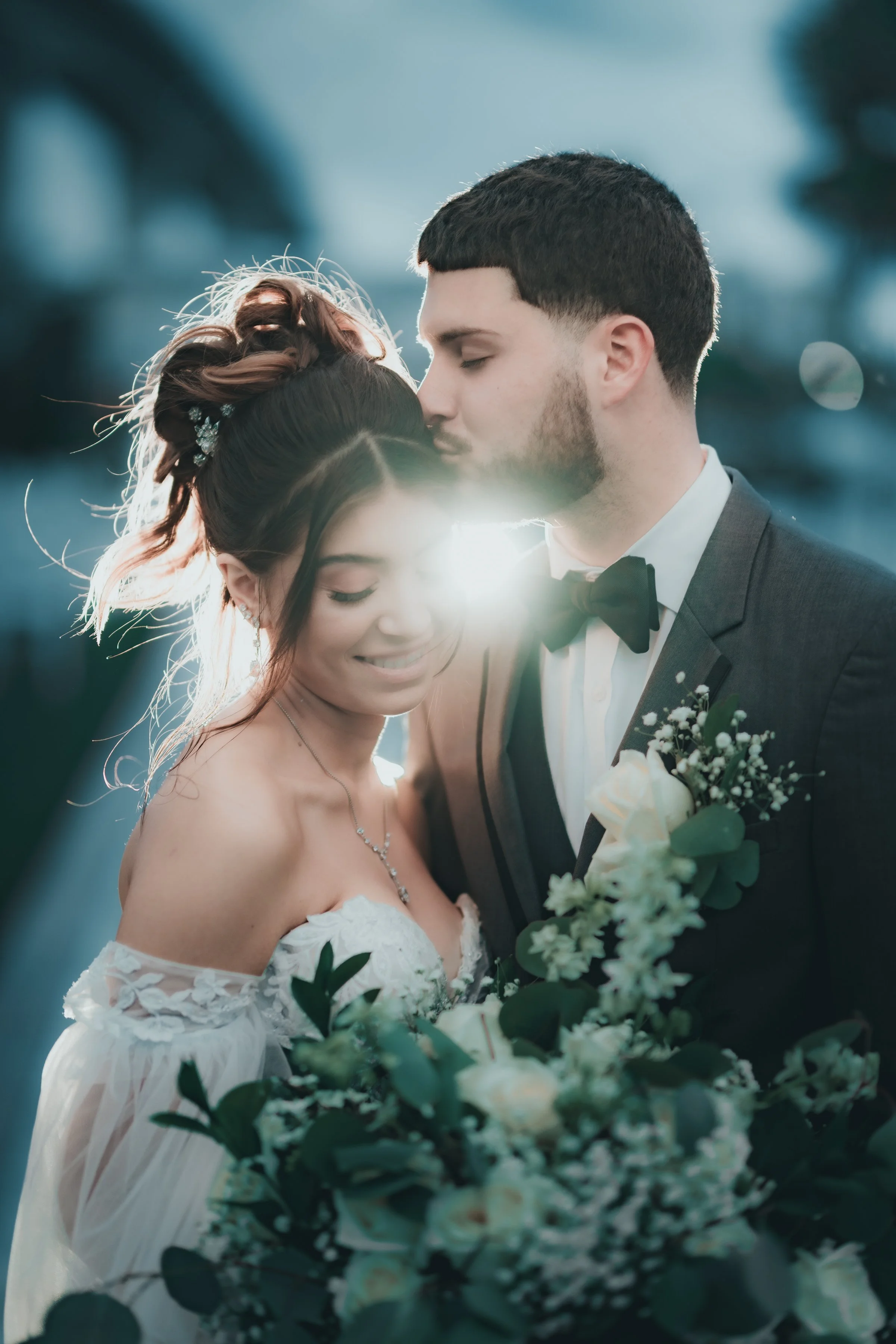 A bride and groom close together outdoors, the groom kissing the bride's forehead, holding a bouquet of white flowers, with sunlight shining behind them.