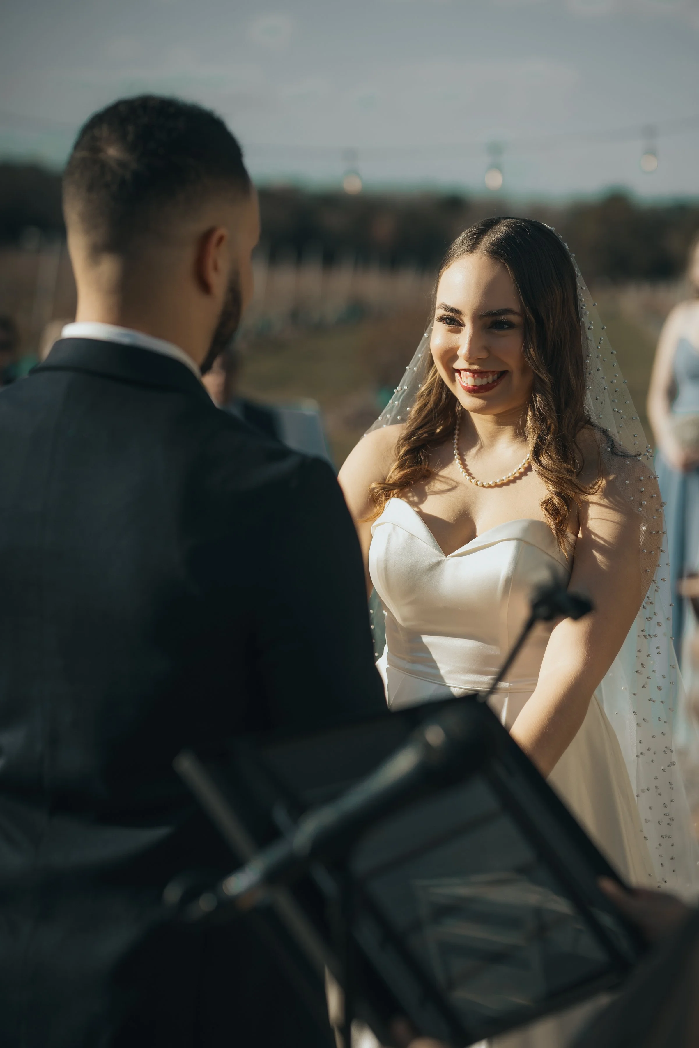 A bride and groom exchanging vows outdoors during their wedding ceremony, with the bride smiling and wearing a white dress and pearl necklace.