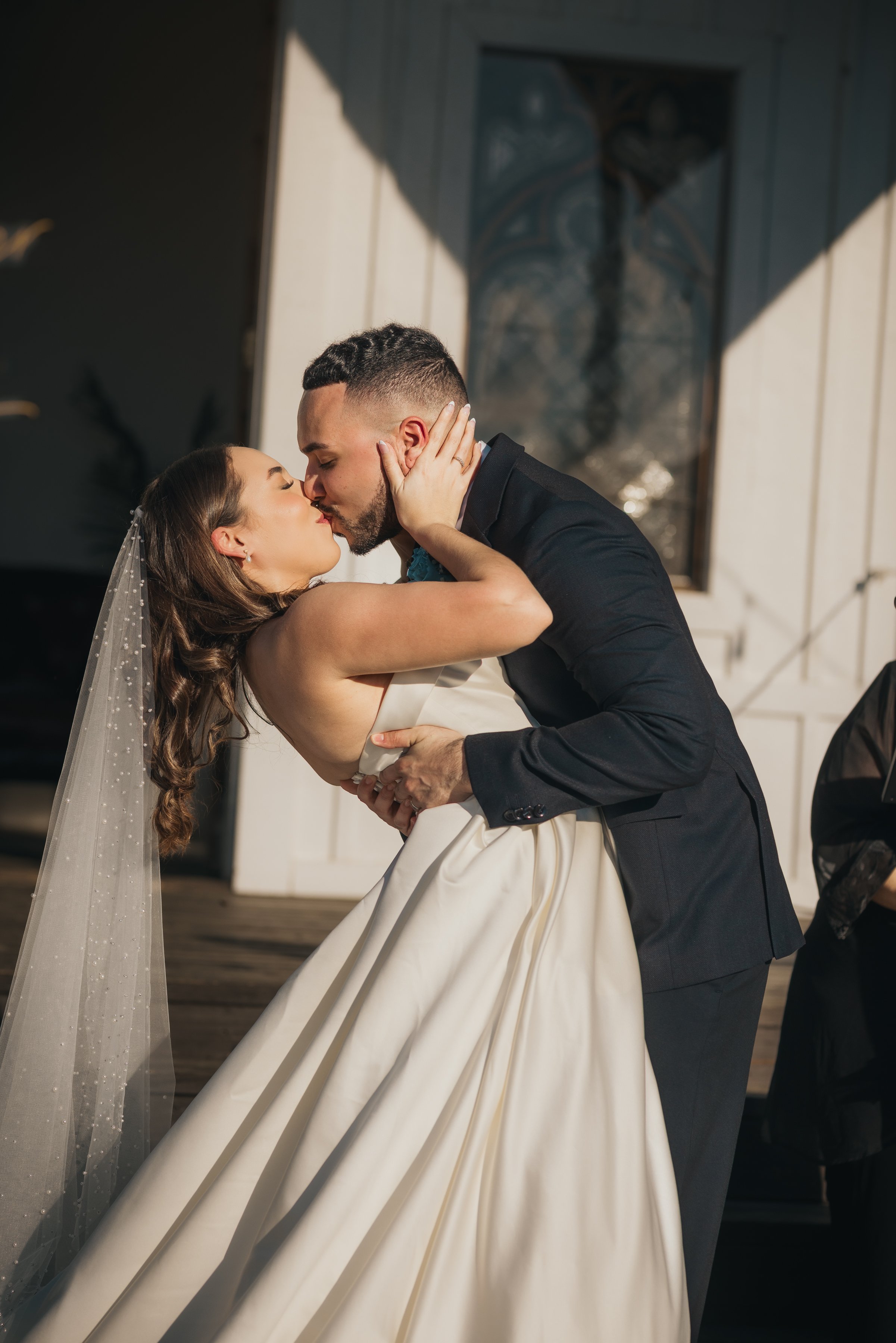 A bride and groom sharing a kiss during their wedding, with the groom dipping the bride forward, outside near a building with a window.