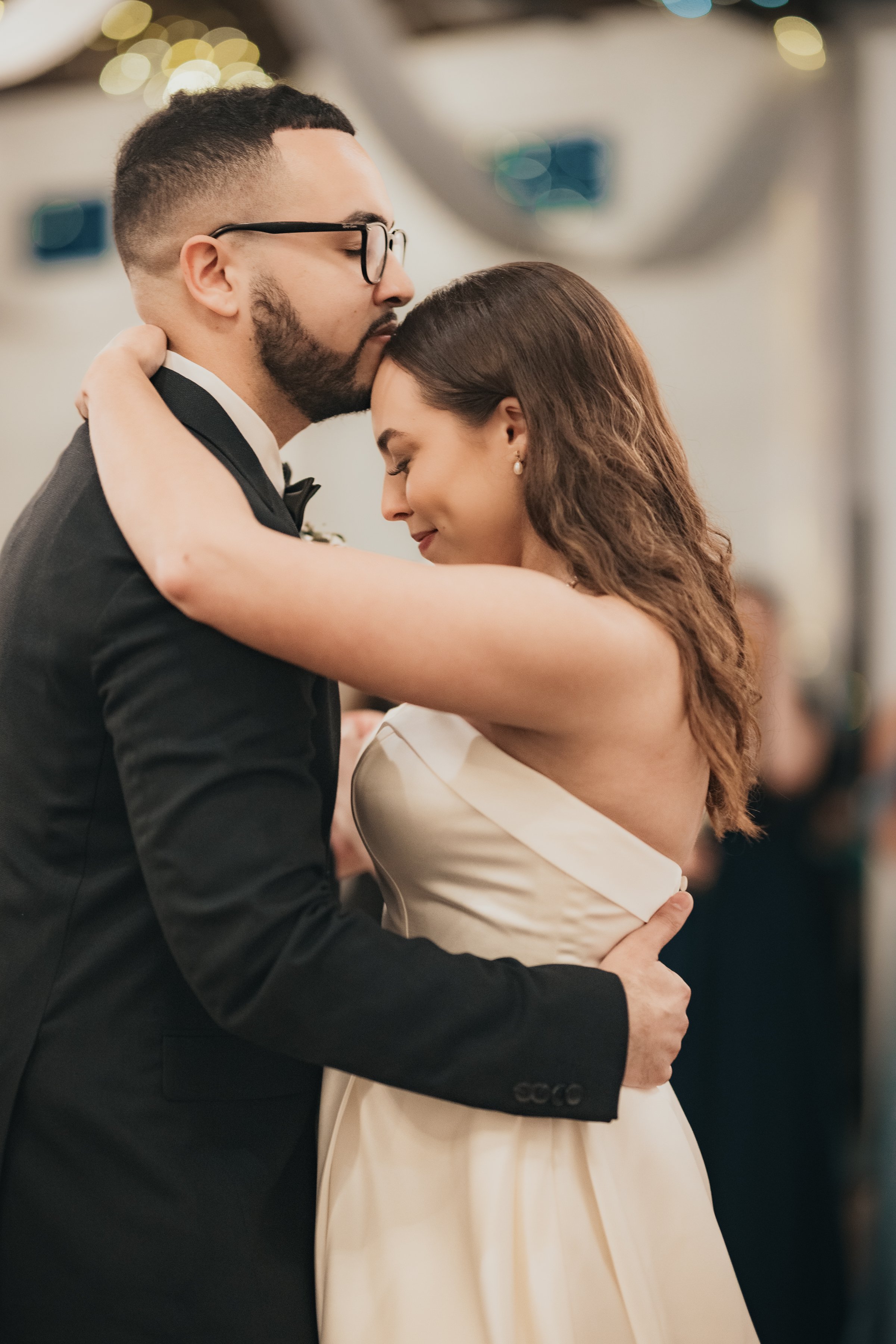 A couple dancing closely at their wedding, with the groom in a black tuxedo and the bride in a strapless white dress, embracing with eyes closed and smiling softly.