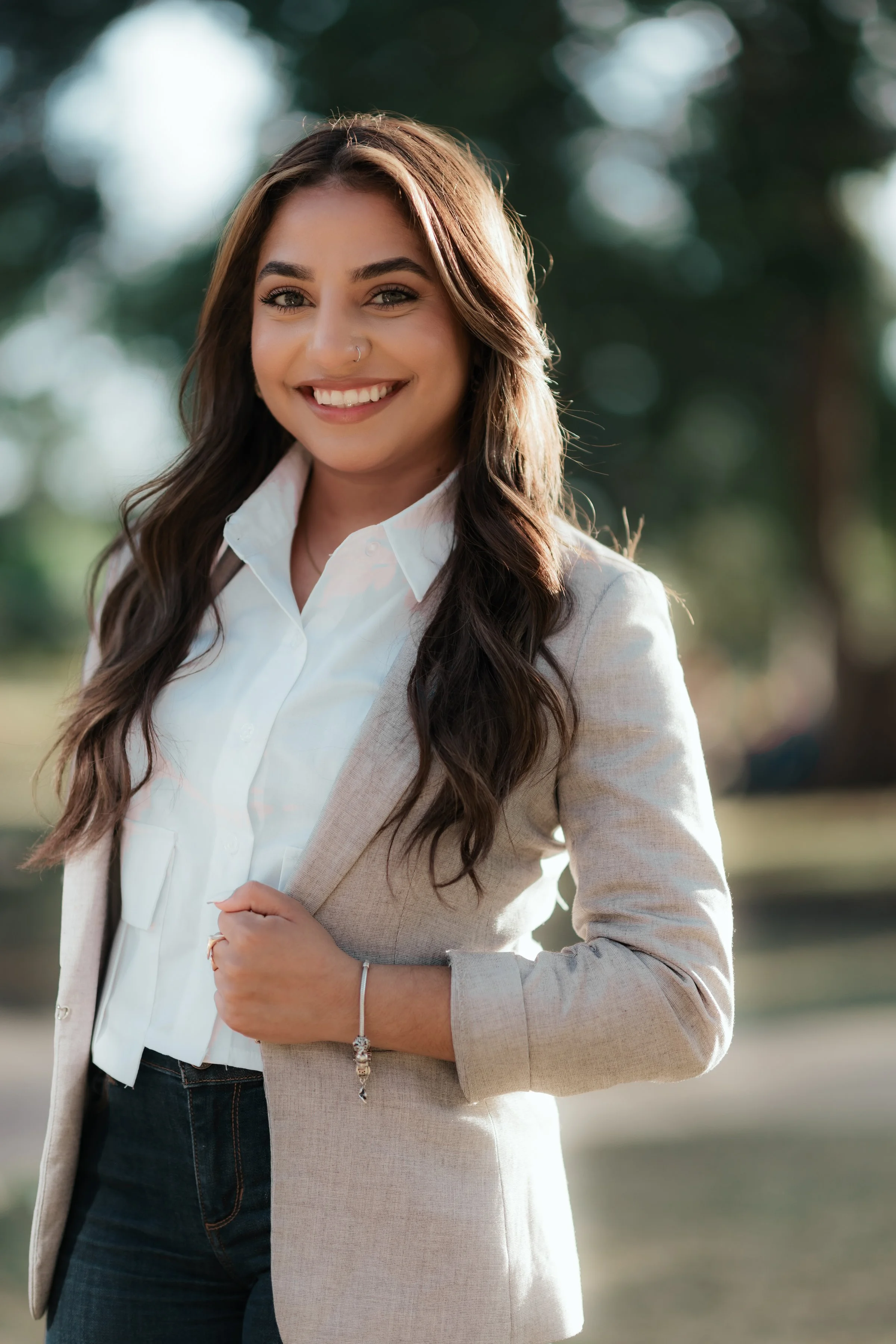 A young woman with long brown hair, smiling, wearing a white shirt, beige blazer, and dark jeans, standing outdoors.