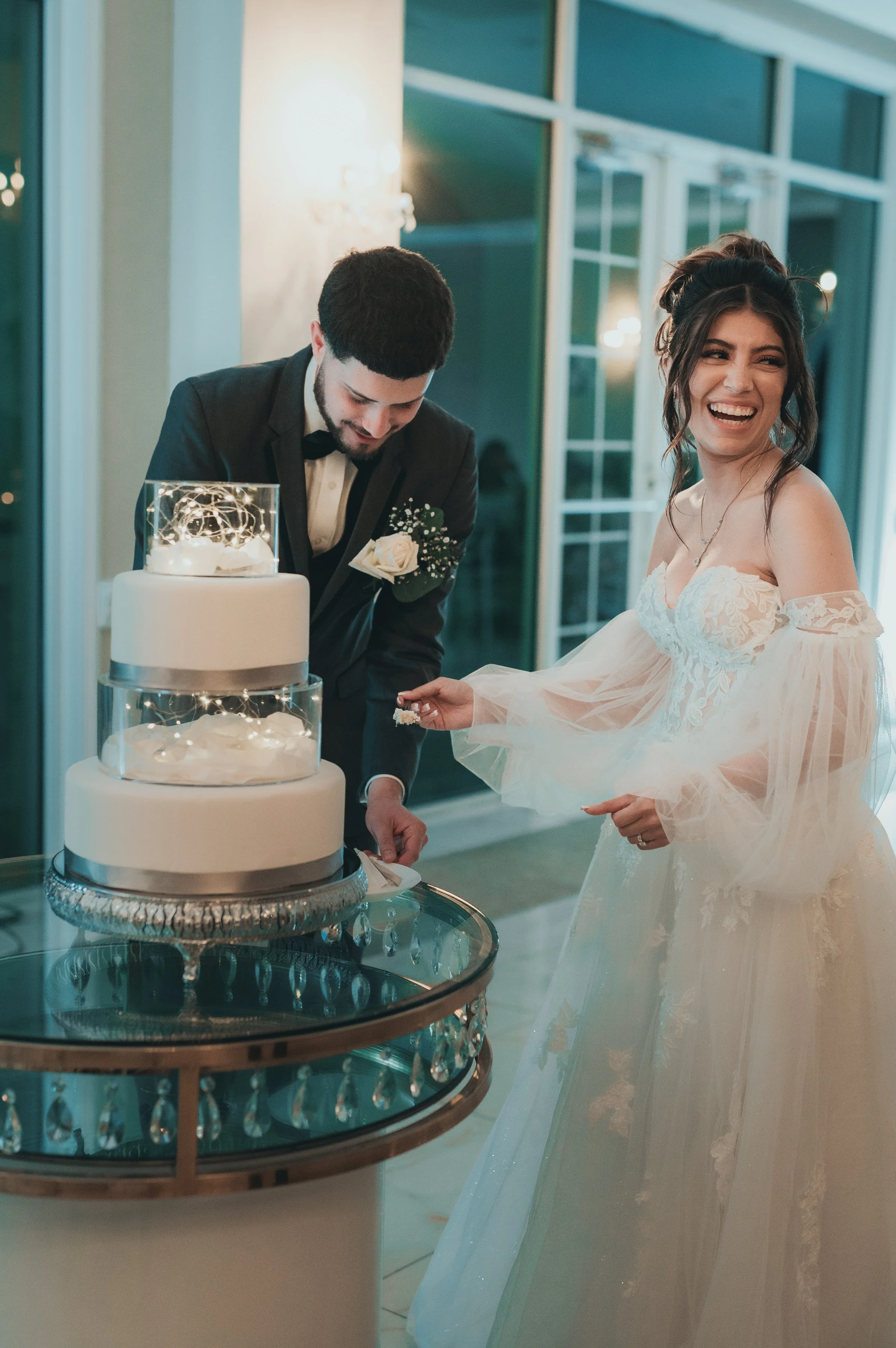A bride and groom cutting a wedding cake together at their reception, both smiling joyfully.