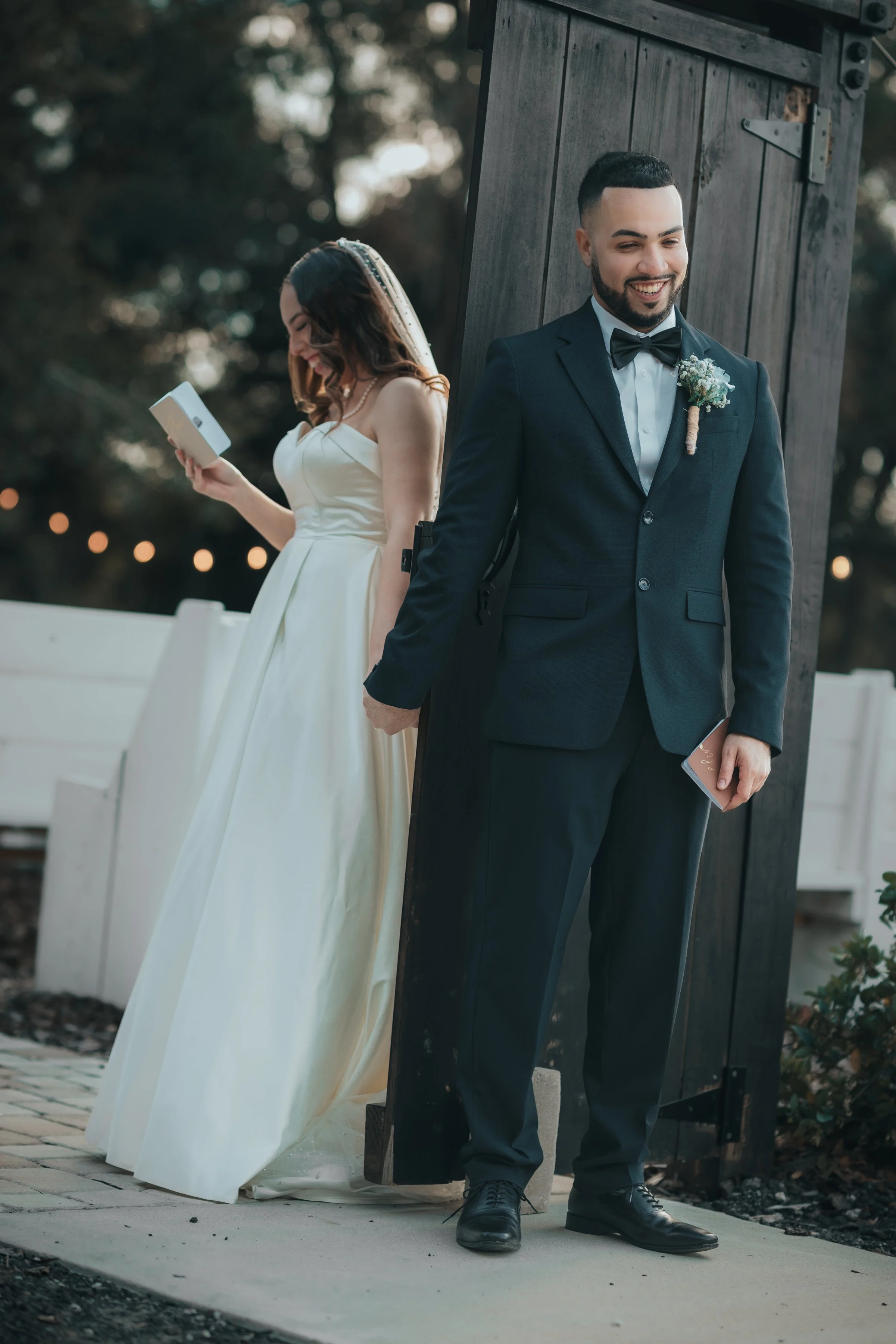 A bride and groom stand back-to-back outdoors during their wedding ceremony. The bride, in a white wedding gown, is looking at her phone and laughing, while the groom, in a navy suit with a bow tie and boutonniere, smiles and also holds his phone.