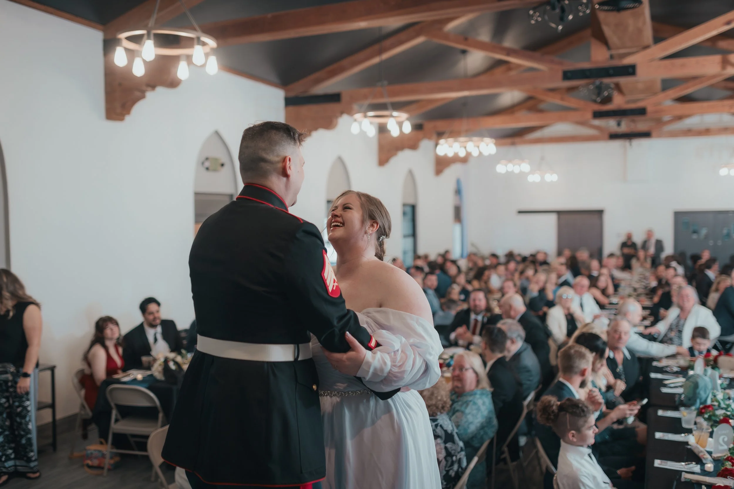 A bride and a groom dancing during a wedding reception, with many guests seated at tables in the background, inside a spacious hall with wooden beams on the ceiling and hanging lights.