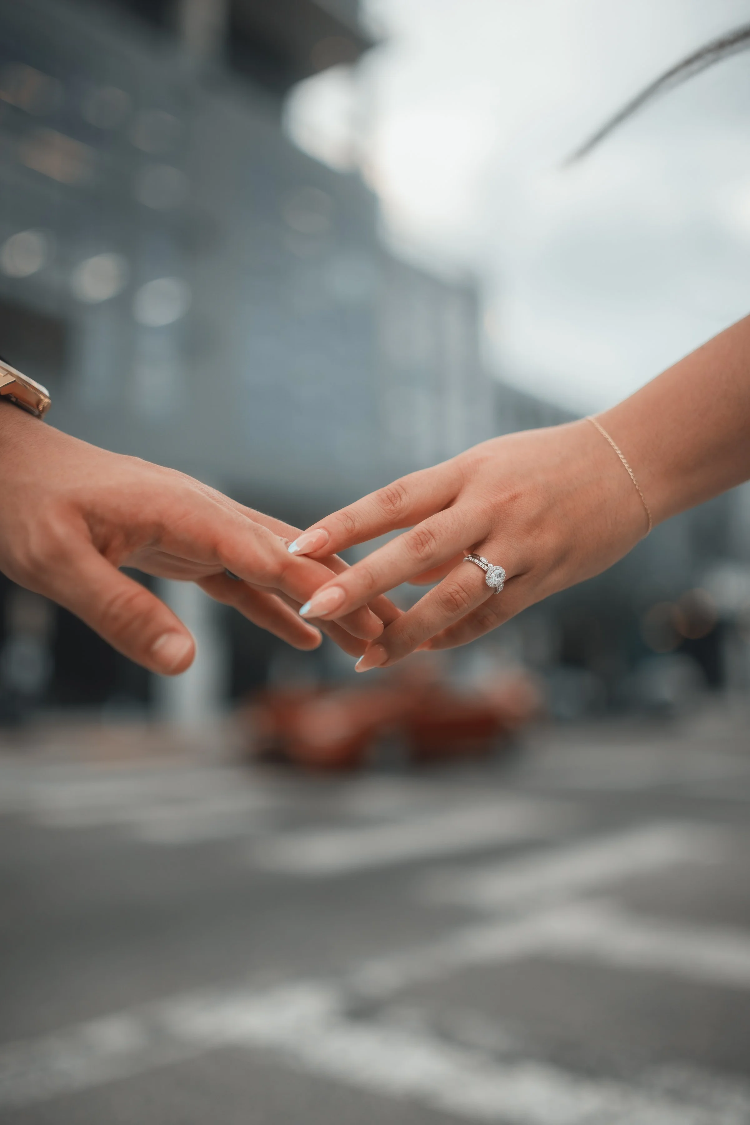 Close-up of two hands reaching to touch, with one hand featuring an engagement ring, set against a blurred city street background.