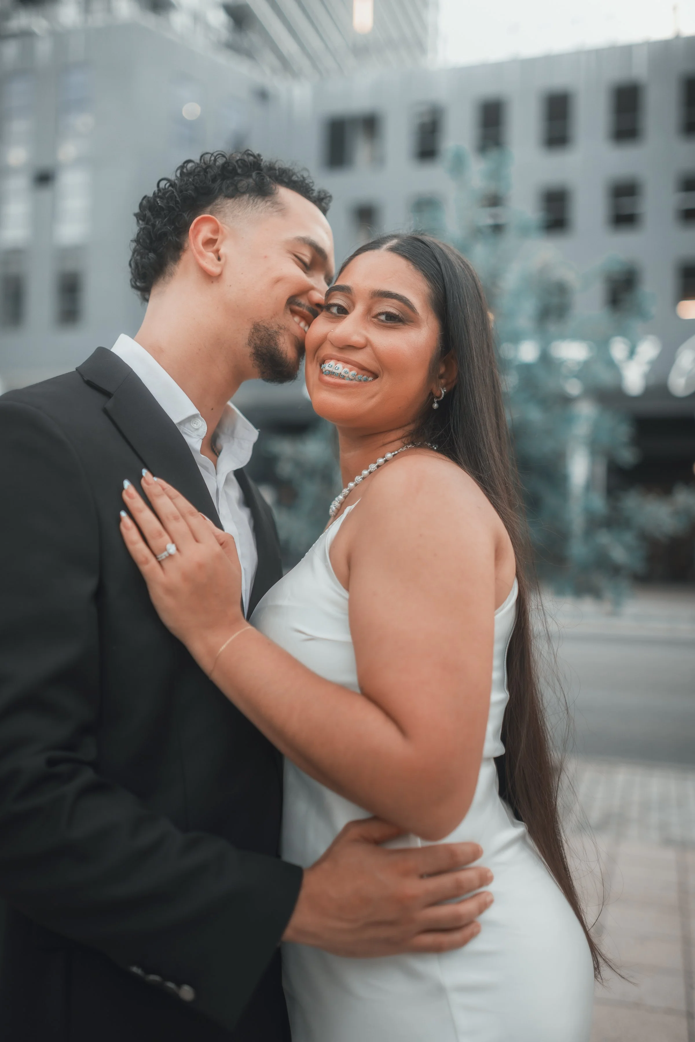 A newlywed couple, a man and a woman, sharing a joyful moment outdoors in an urban setting. The man is wearing a black suit, and the woman is in a white dress with jewelry. They are close, smiling, and showing affection.