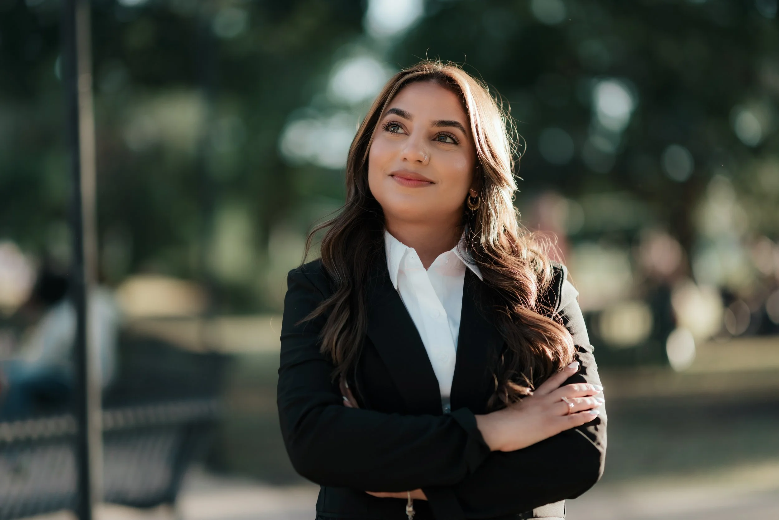 A young woman with long wavy brown hair, wearing a black blazer and white shirt, standing outdoors with her arms crossed, smiling and looking to the side.
