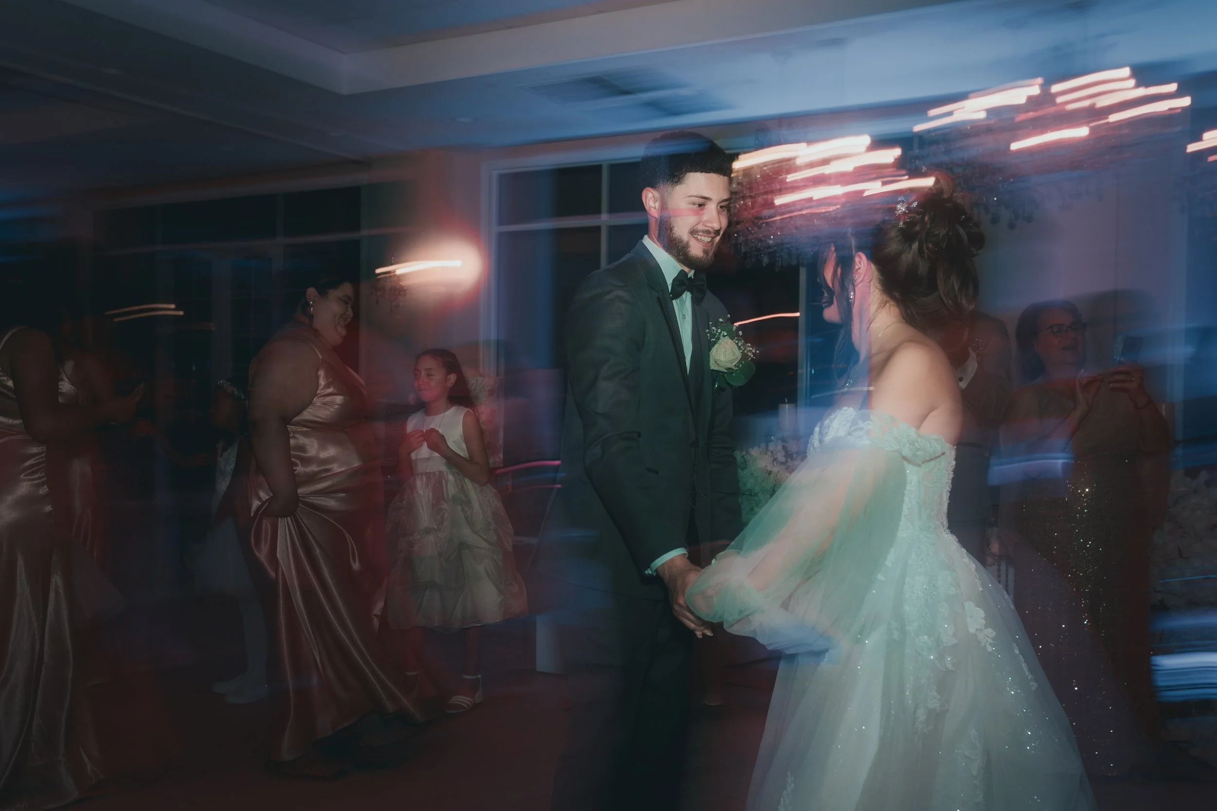 A bride and groom dancing at their wedding reception, surrounded by guests. The scene is blurred with streaks of light, indicating motion and a lively atmosphere.