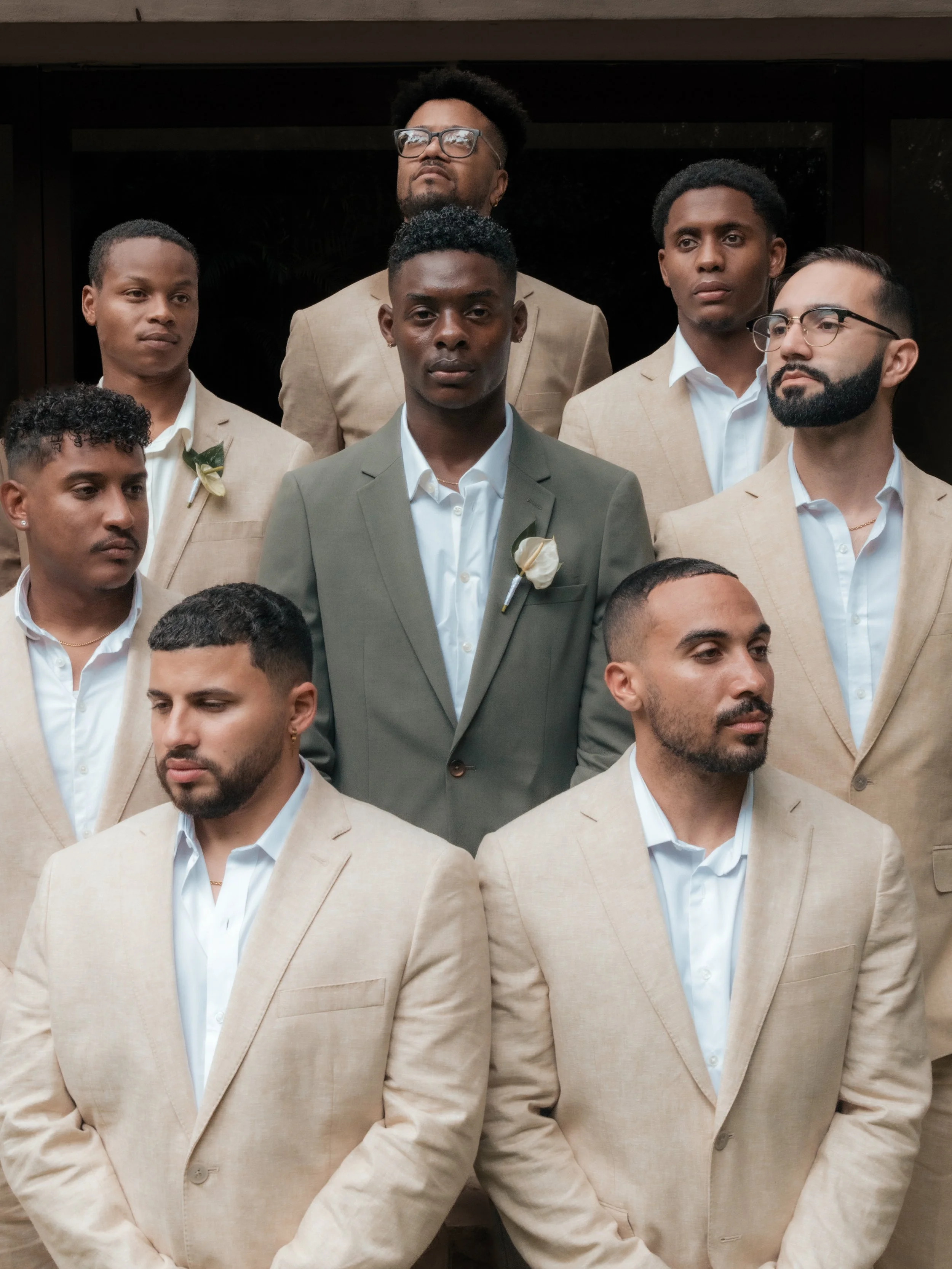 A group of nine men dressed in formal suits, with the front row wearing beige suits and the middle man in a gray suit, standing outdoors in front of a dark background.