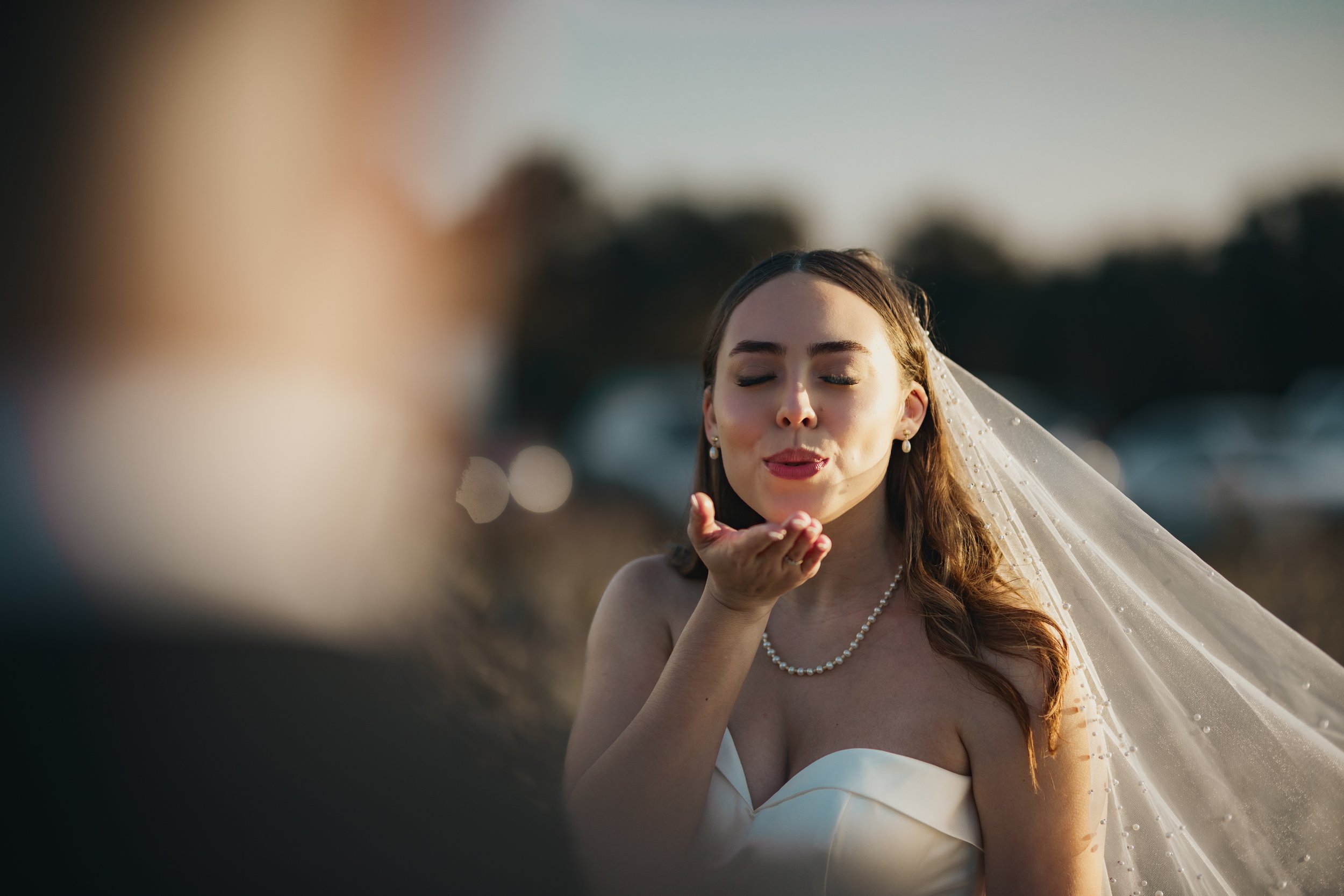Bride with long brown hair, wearing a white strapless wedding gown and a pearl necklace, blowing a kiss outdoors during sunset.