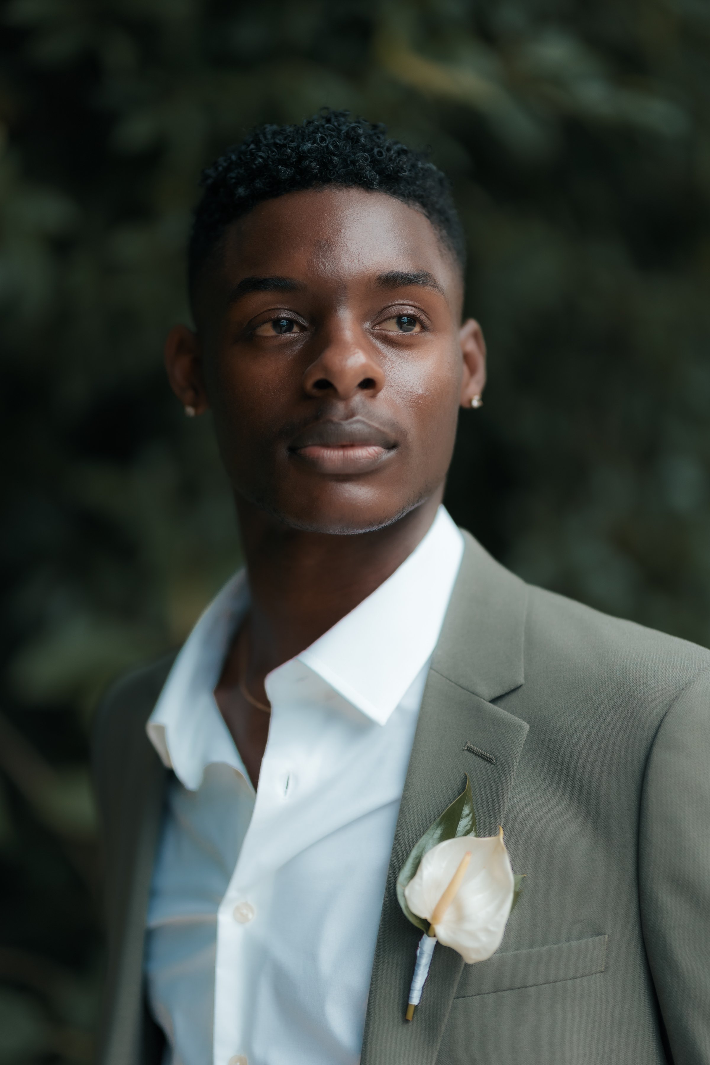 Portrait of a young man in a suit with a white flower boutonniere, standing outdoors with dark green foliage background.