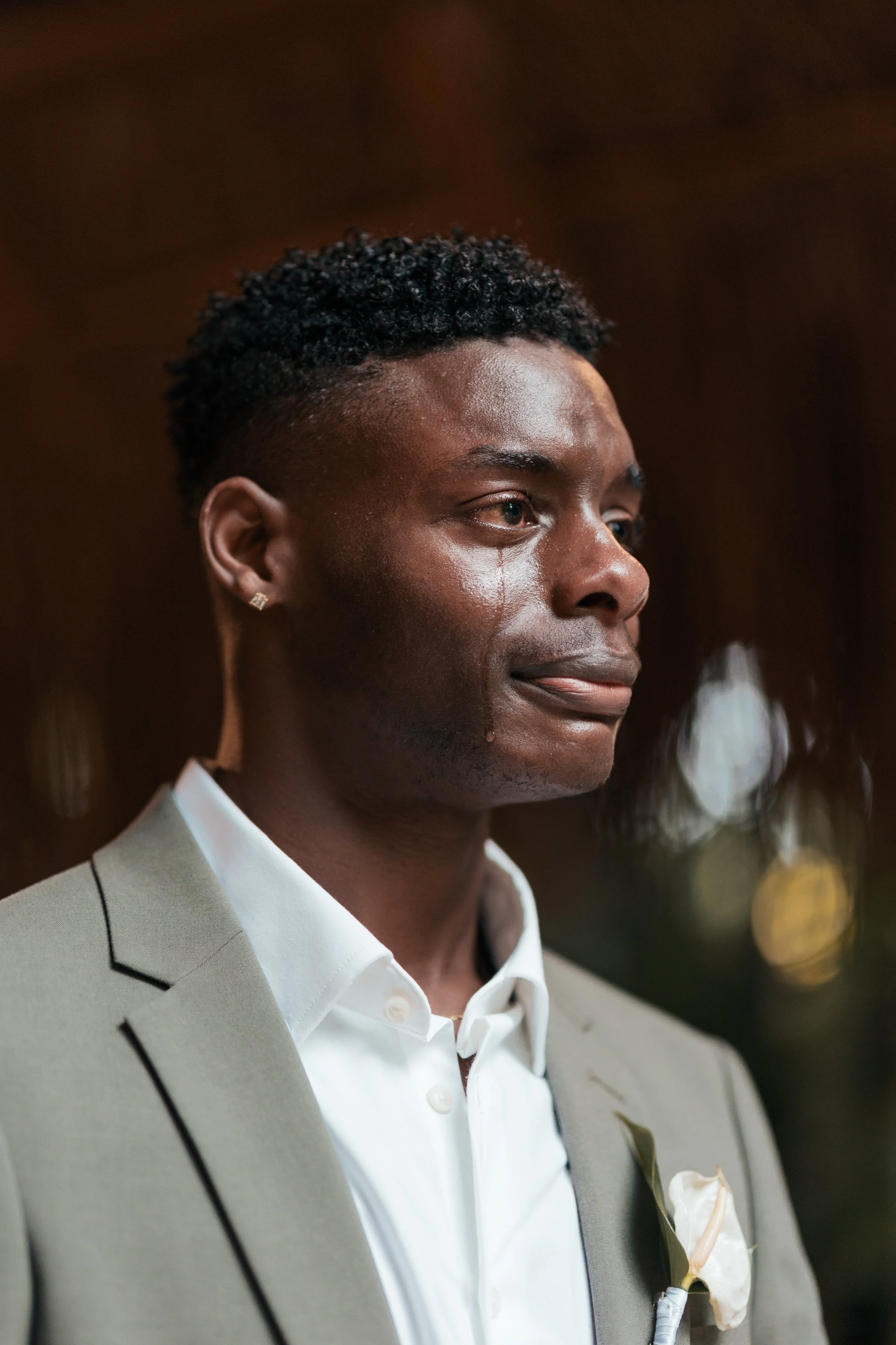 A man with dark curly hair, wearing a light gray suit and a white shirt, with a boutonniere, looks contemplative with a tear in his eye.