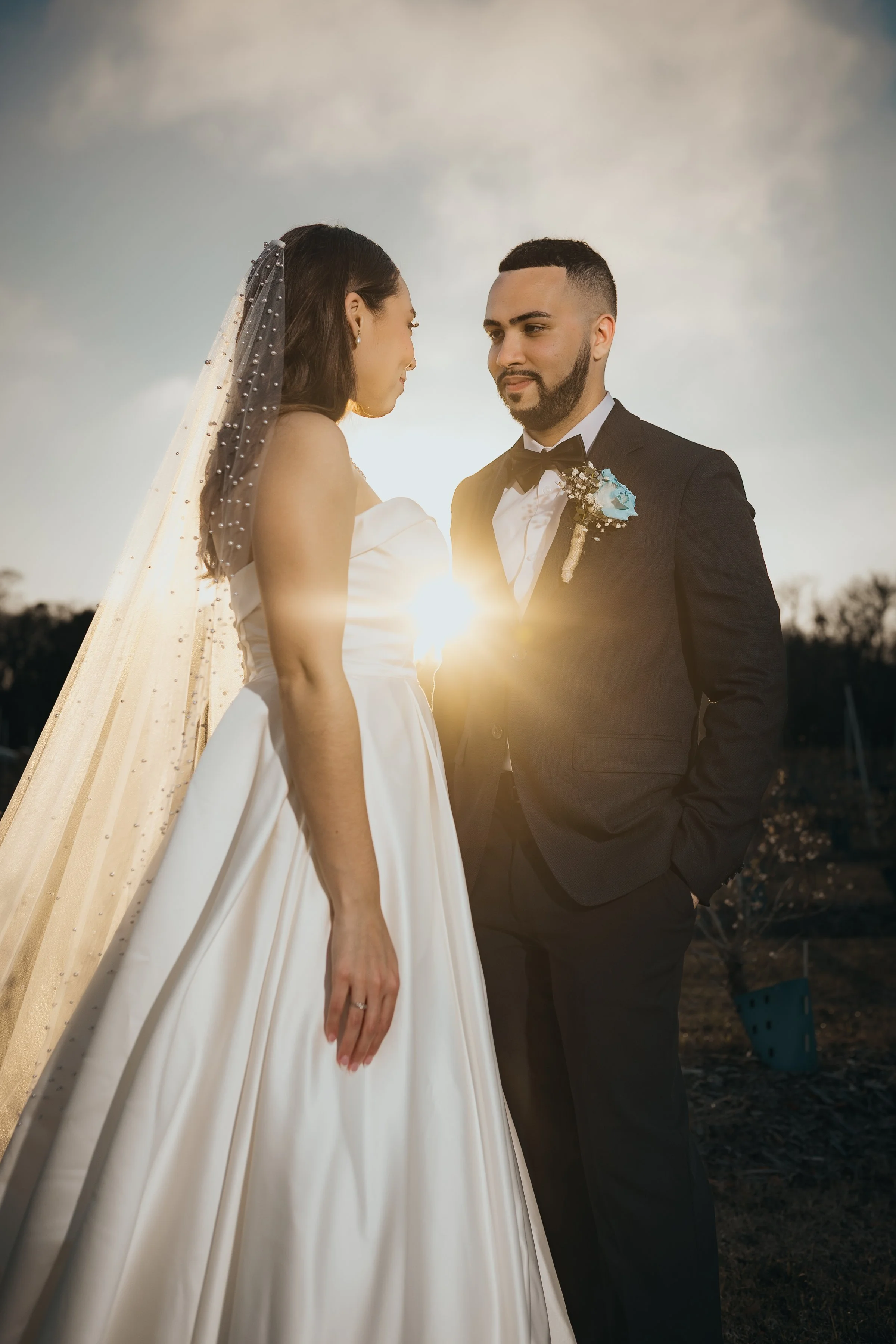 A bride and groom standing face to face outdoors during sunset, with the sun behind them creating a warm glow, dressed in wedding attire.