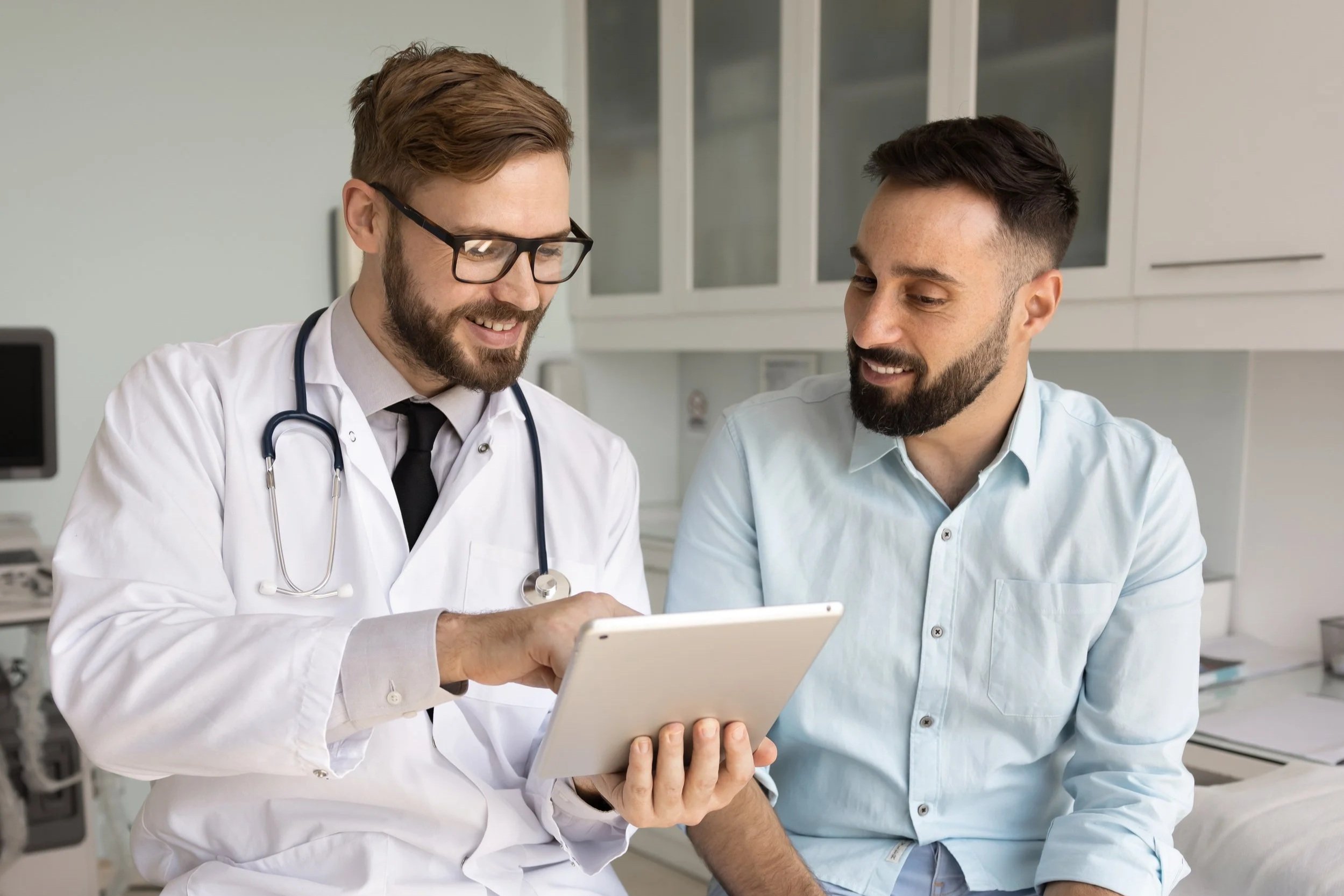Digital modernization makes work easier for professionals, and here two men, one in a doctor's coat with a stethoscope and glasses, and another in a light blue shirt, are looking at a tablet together in a clinical setting.