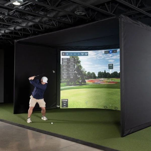 Person practicing golf swing inside indoor golf simulator with a large screen displaying a golf course.