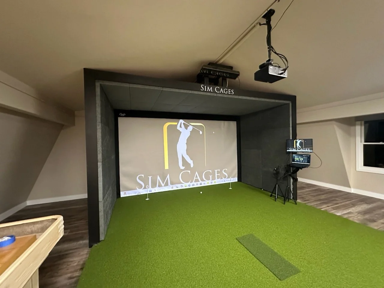 Indoor golf simulation room with green turf, a large screen displaying the logo 'S.I.M CAGES' and an illustration of a golfer, and equipment including a monitor and projector, with a sign labeled 'SIM CAGES' above the setup.