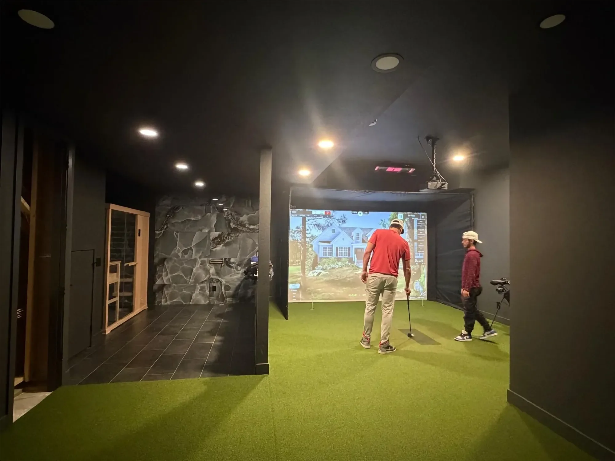 Indoor golf simulation room with a large screen displaying a virtual golf course, two men preparing to golf, artificial grass floor, and a wall with stone decor.