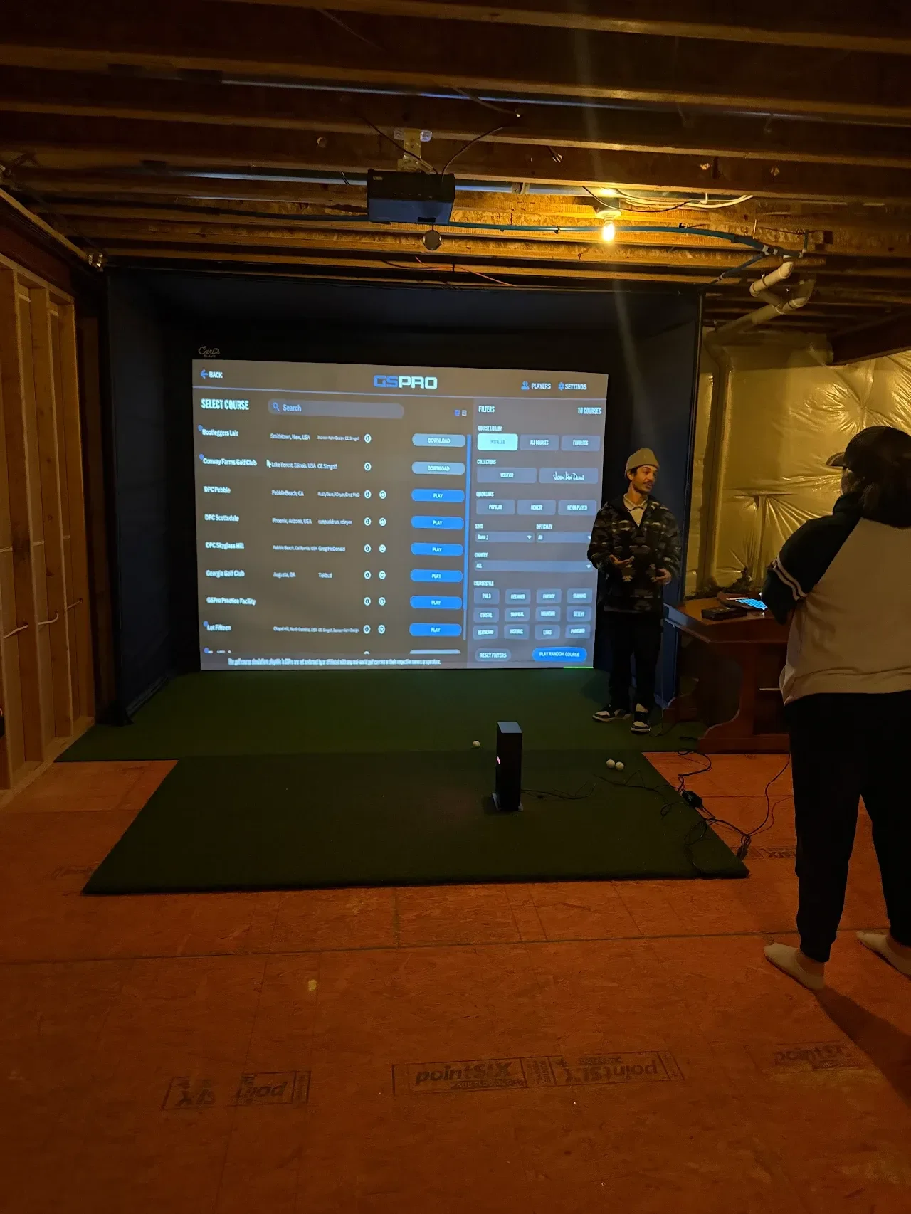Indoor golf simulation setup with a large screen displaying a course selection menu, artificial grass hitting area, and two people standing and interacting with the system.