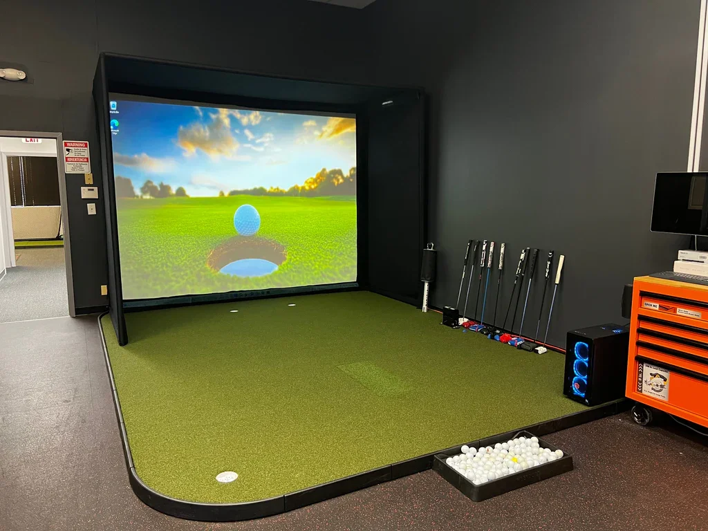 Indoor golf simulator with a large screen showing a golf course, golf clubs on a rack, golf balls in a tray, and a projector on a rolling cart.