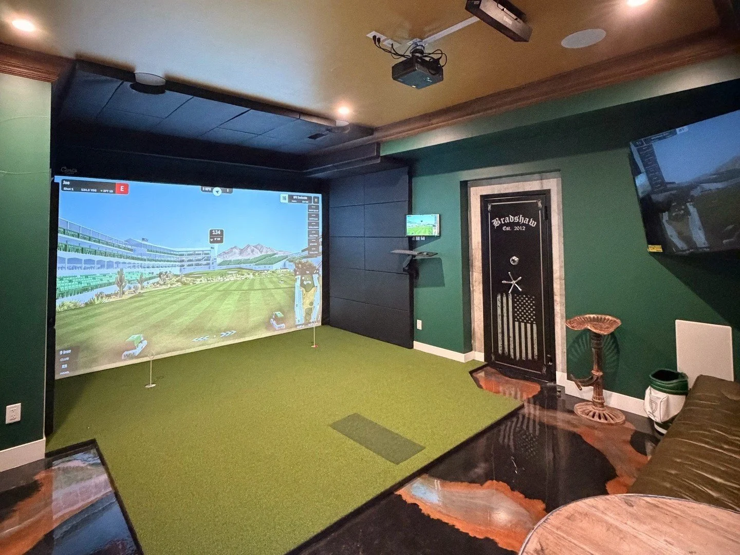 Indoor golf simulator room with a large screen displaying a golf course, green carpeted area, wall-mounted TV, black door with American flag design, and wooden furniture.