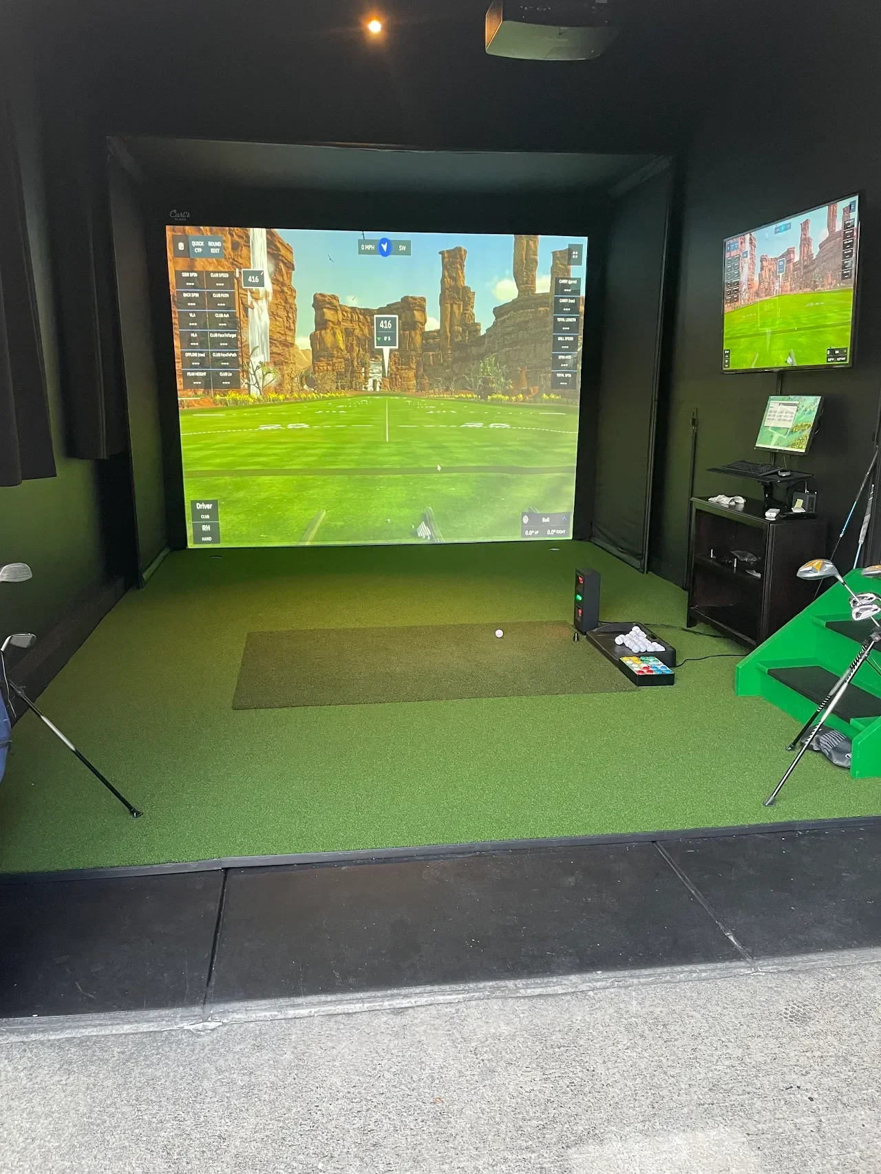 Indoor golf simulation room with a large screen showing a virtual golf course, golf clubs, and accessories on the floor, along with additional monitors on the wall.