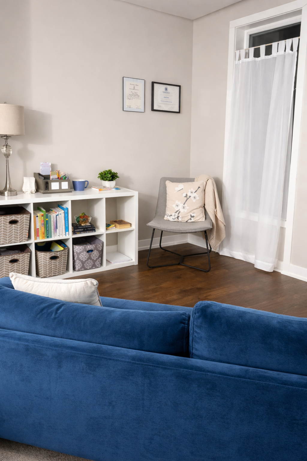 A cozy living room corner with a blue sofa, a white cube storage unit with books and storage baskets, a gray chair with a floral cushion, a white curtain, a wooden floor, and a wall with framed certificates.