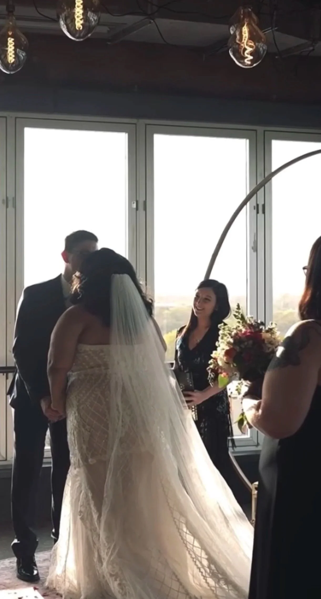 A wedding ceremony with a bride and groom holding hands in front of an officiant, with a bridesmaid holding a bouquet of flowers nearby.